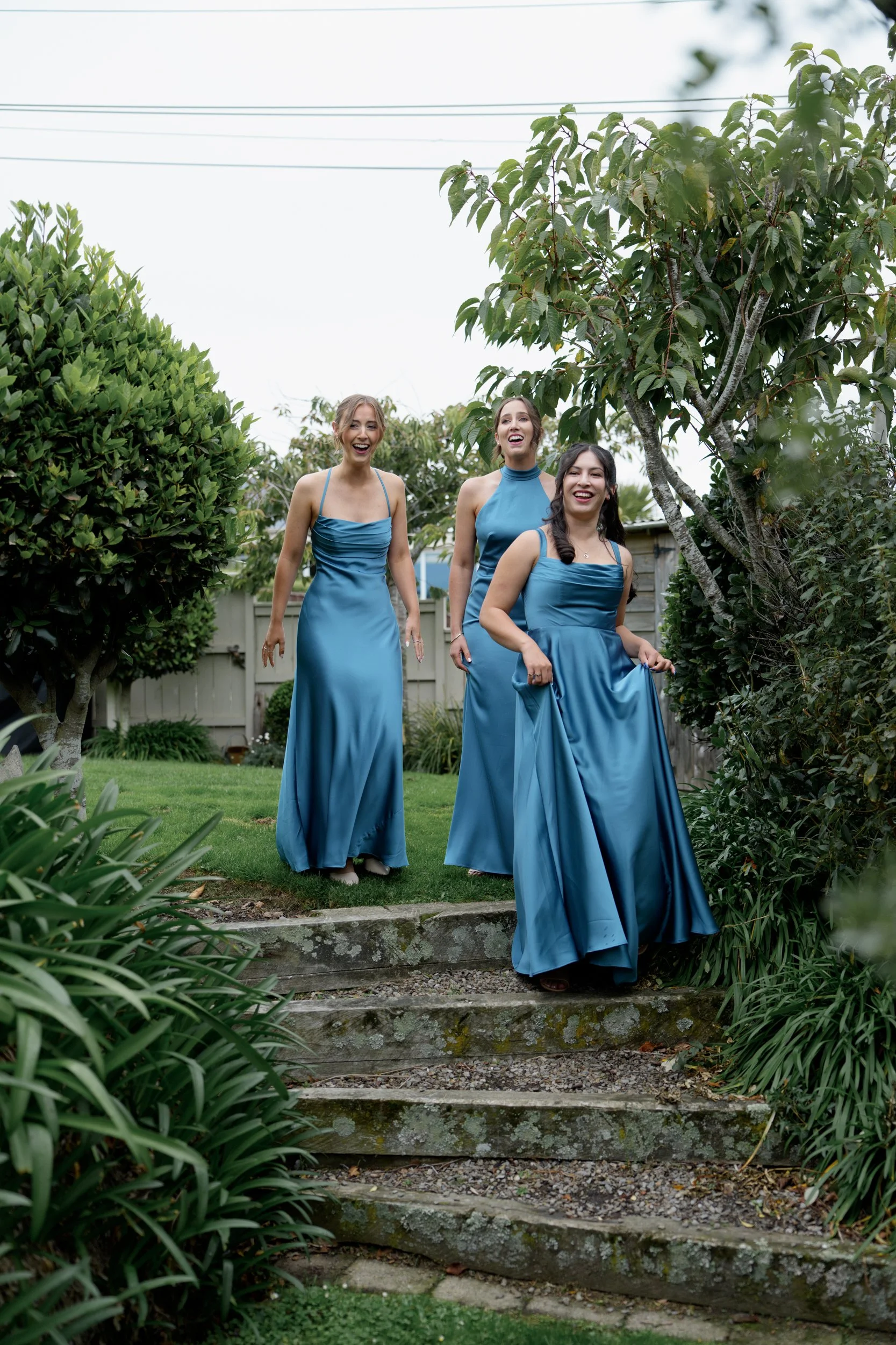 Film + Digital Wedding Photographer Dunedin - Lochend Woolshed, Dunedin, Otago, New Zealand. Three bridesmaids in matching blue satin gowns are walking outdoors down a small stone staircase, surrounded by green bushes and trees, and smiling happily.