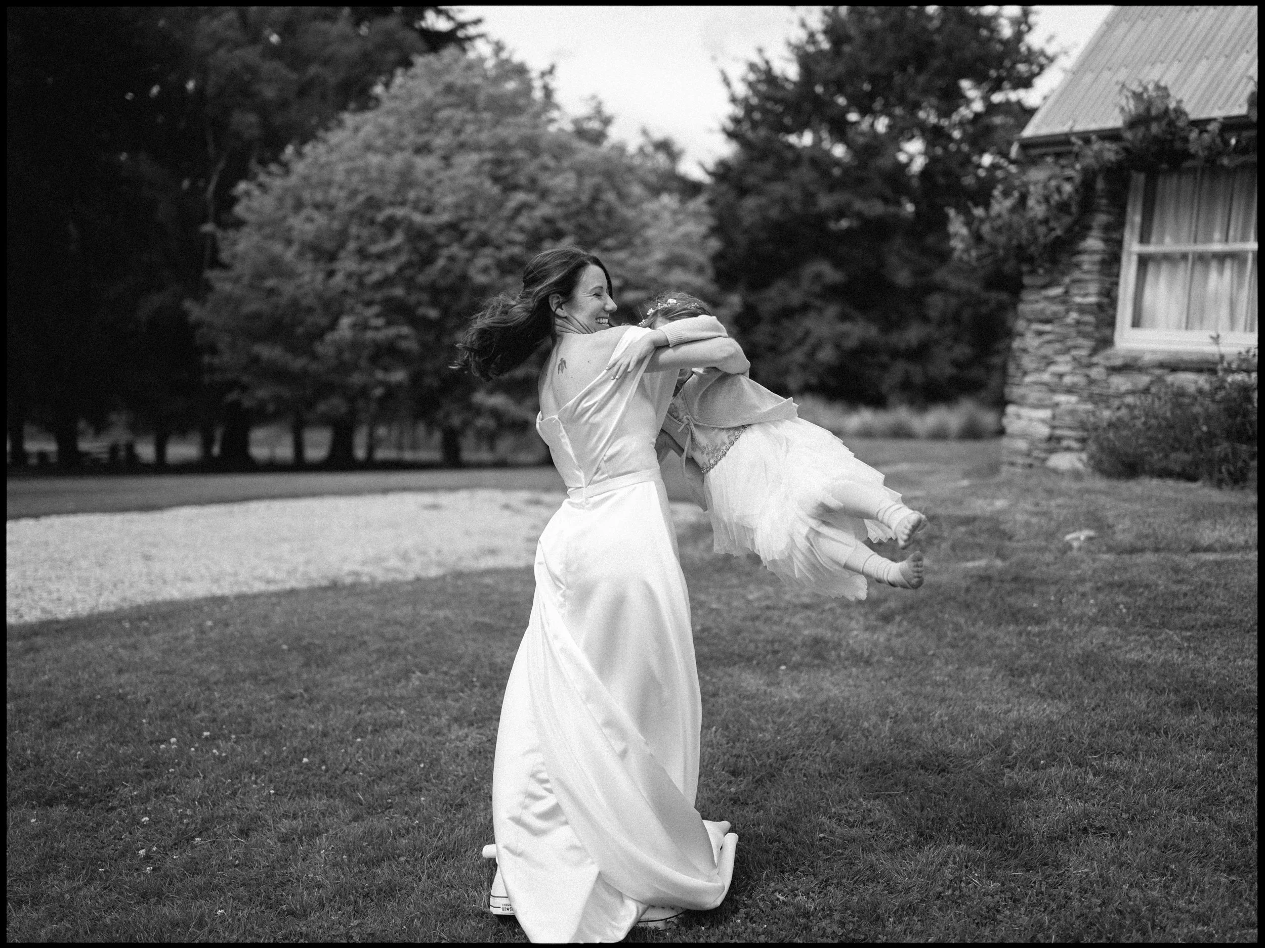 Queenstown Elopement, Otago, New Zealand. Bride in a wedding dress lifting and spinning her young daughter in a white dress outdoors, with trees and a house in the background.