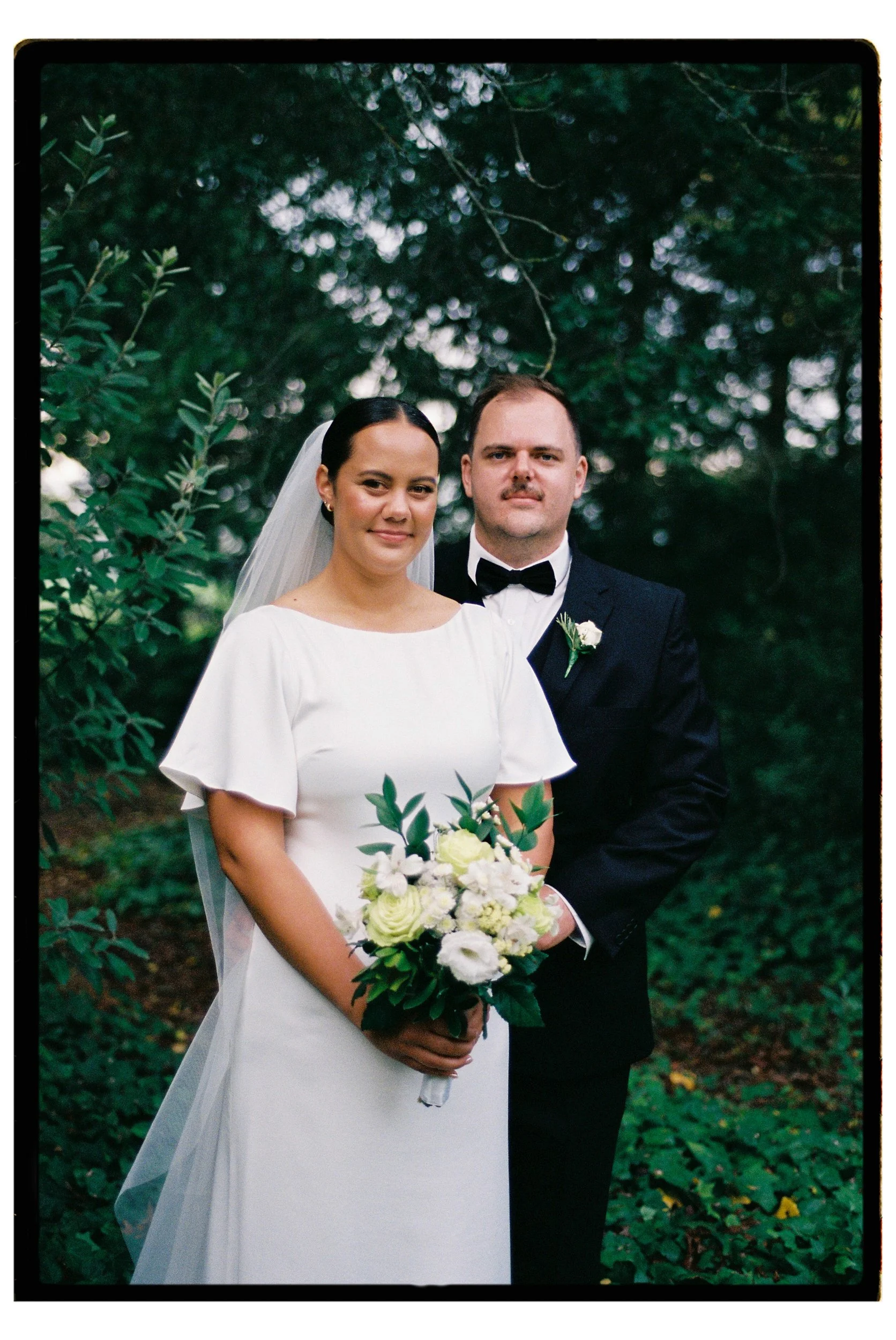 Film + Digital Wedding Photographer Christchurch - Backyard Wedding, Christchurch, Canterbury, New Zealand. Film photo of bride and groom standing together outdoors surrounded by greenery, dressed in wedding attire.