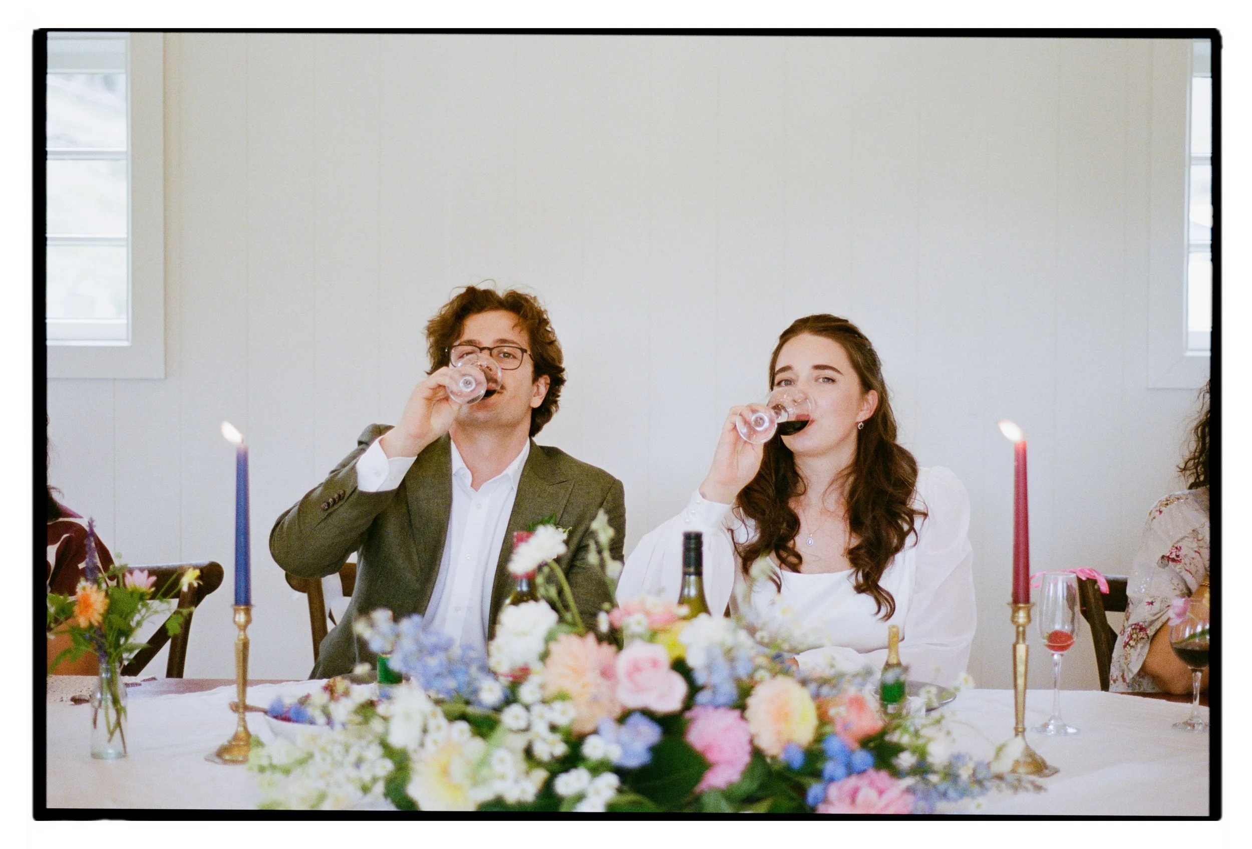 Film + Digital Wedding Photographer Christchurch - Accrington Farmhouse Wedding, Banks Peninsula, Christchurch, New Zealand. Film photo of bride and groom sitting at the dinner table drinking wine. Table is set with colourful candles and flowers.