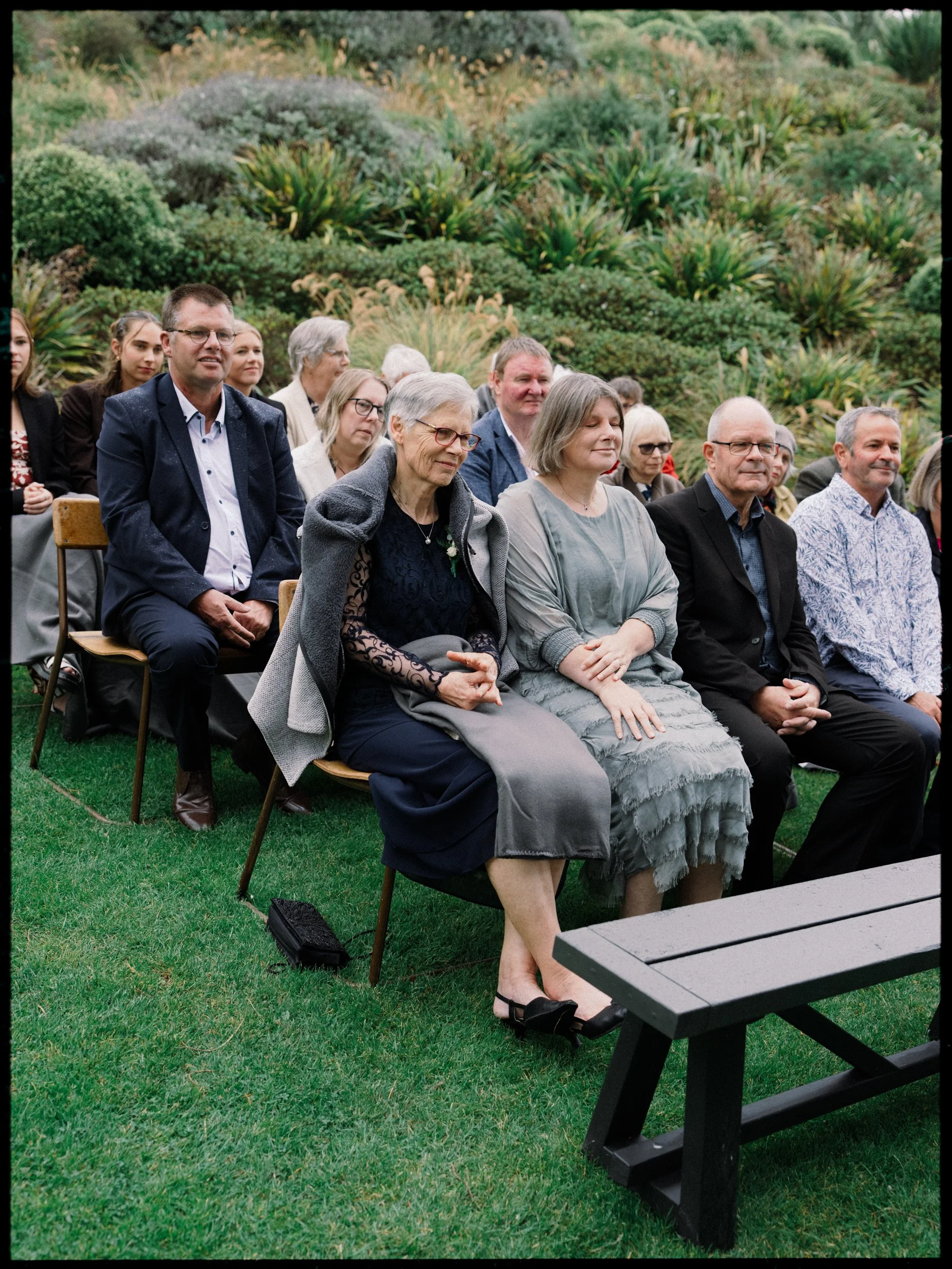 Film + Digital Wedding Photographer Dunedin - Lochend Woolshed, Dunedin, Otago, New Zealand. Guests seated outdoors at a rainy garden wedding ceremony with lush green plants and bushes in the background.