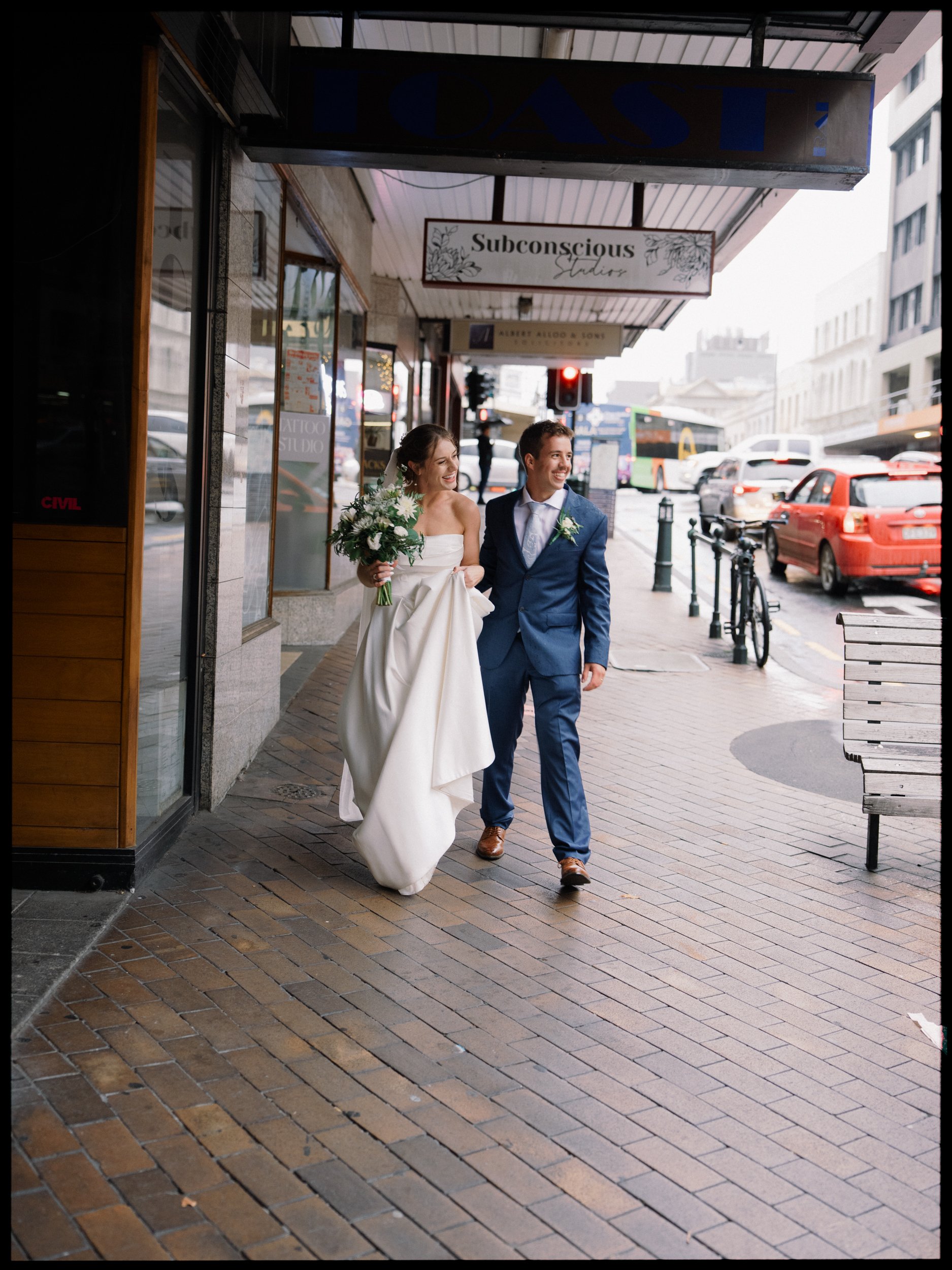 Film + Digital Wedding Photographer Dunedin - Lochend Woolshed, Dunedin, Otago, New Zealand. Bride in white wedding dress and groom in blue suit walking on city sidewalk, smiling and holding hands.