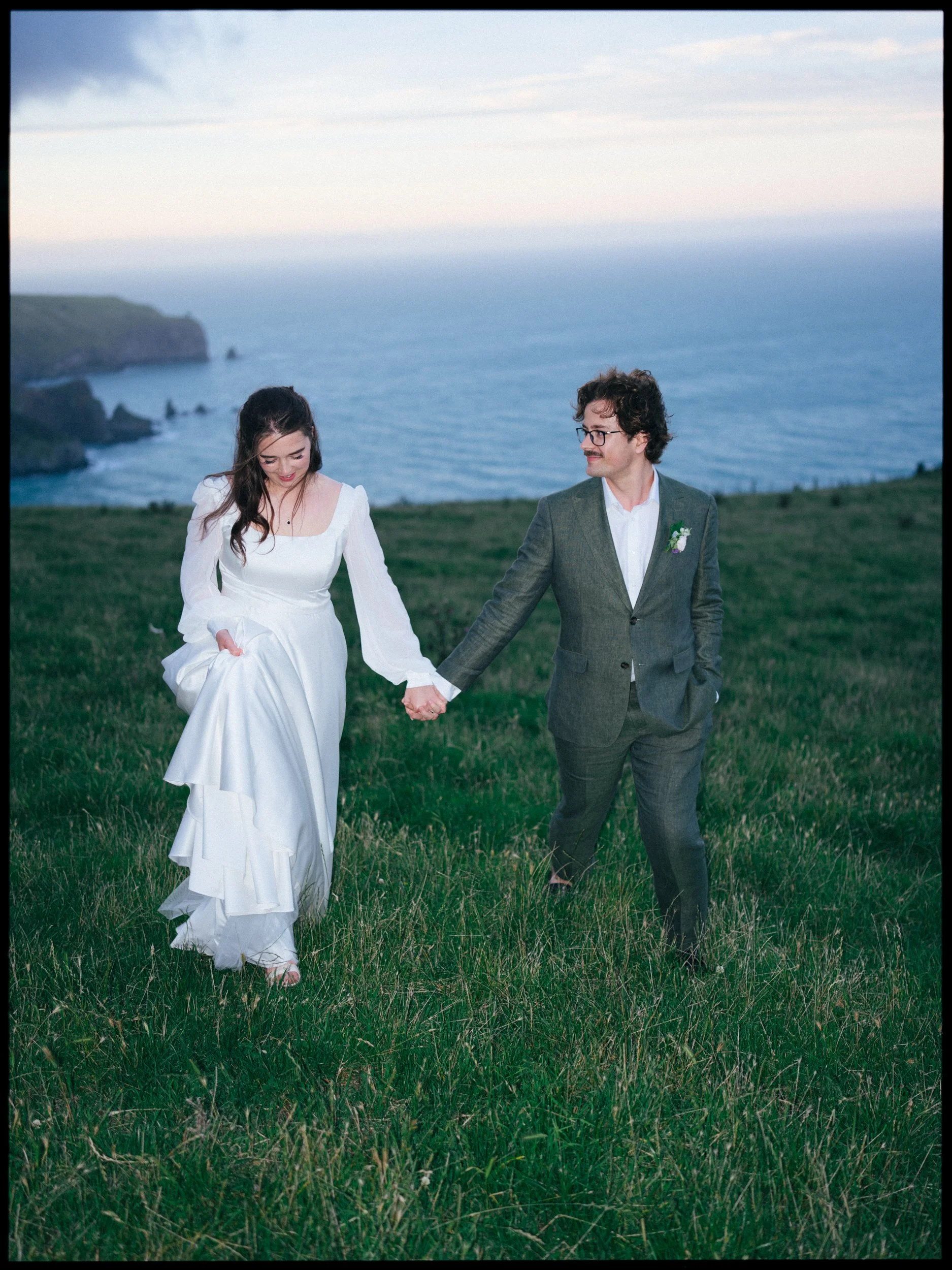 Accrington Farmhouse Wedding, Banks Peninsula, Christchurch, New Zealand. Bride and groom holding hands and walking through a grassy field near the coast, with cliffs and the ocean in the background at sunset.