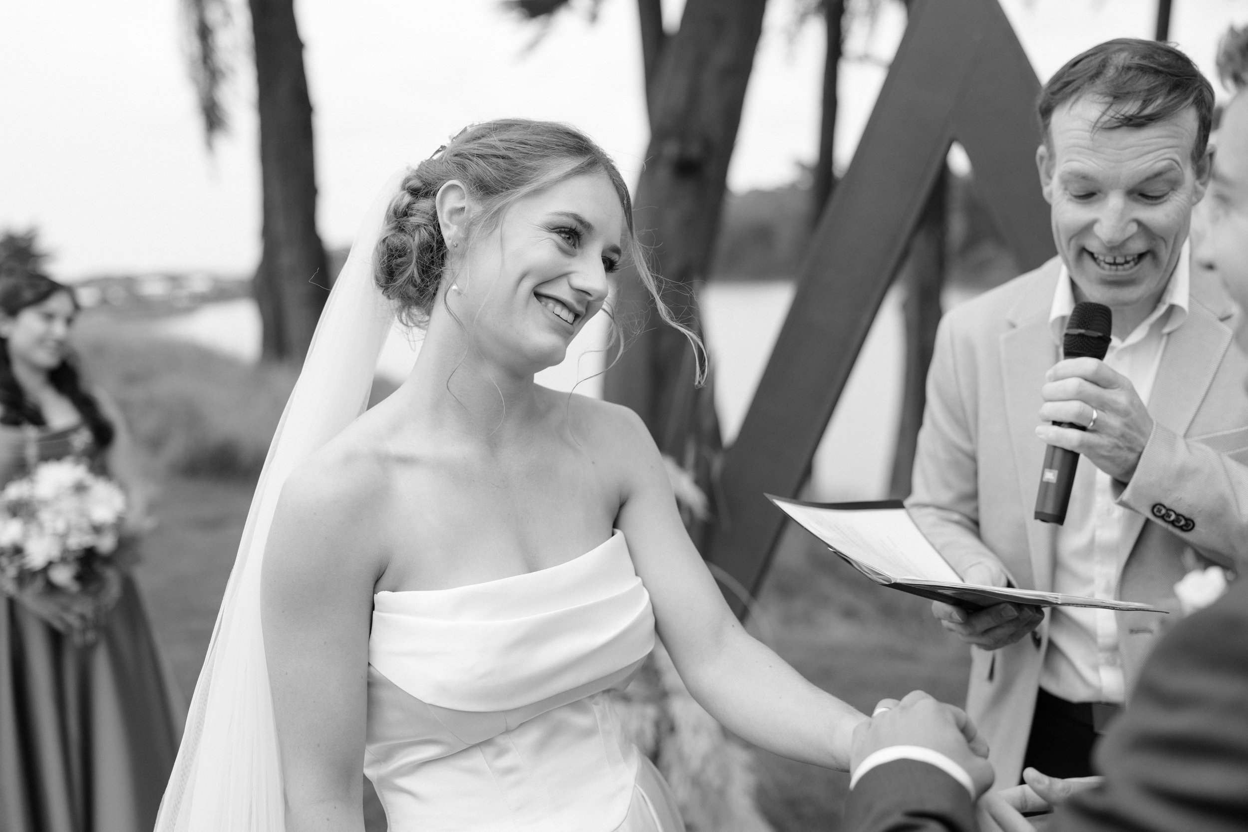 Film + Digital Wedding Photographer Dunedin - Lochend Woolshed, Dunedin, Otago, New Zealand. Bride and groom holding hands during their wedding ceremony outdoors, with celebrant officiating and guests in the background.