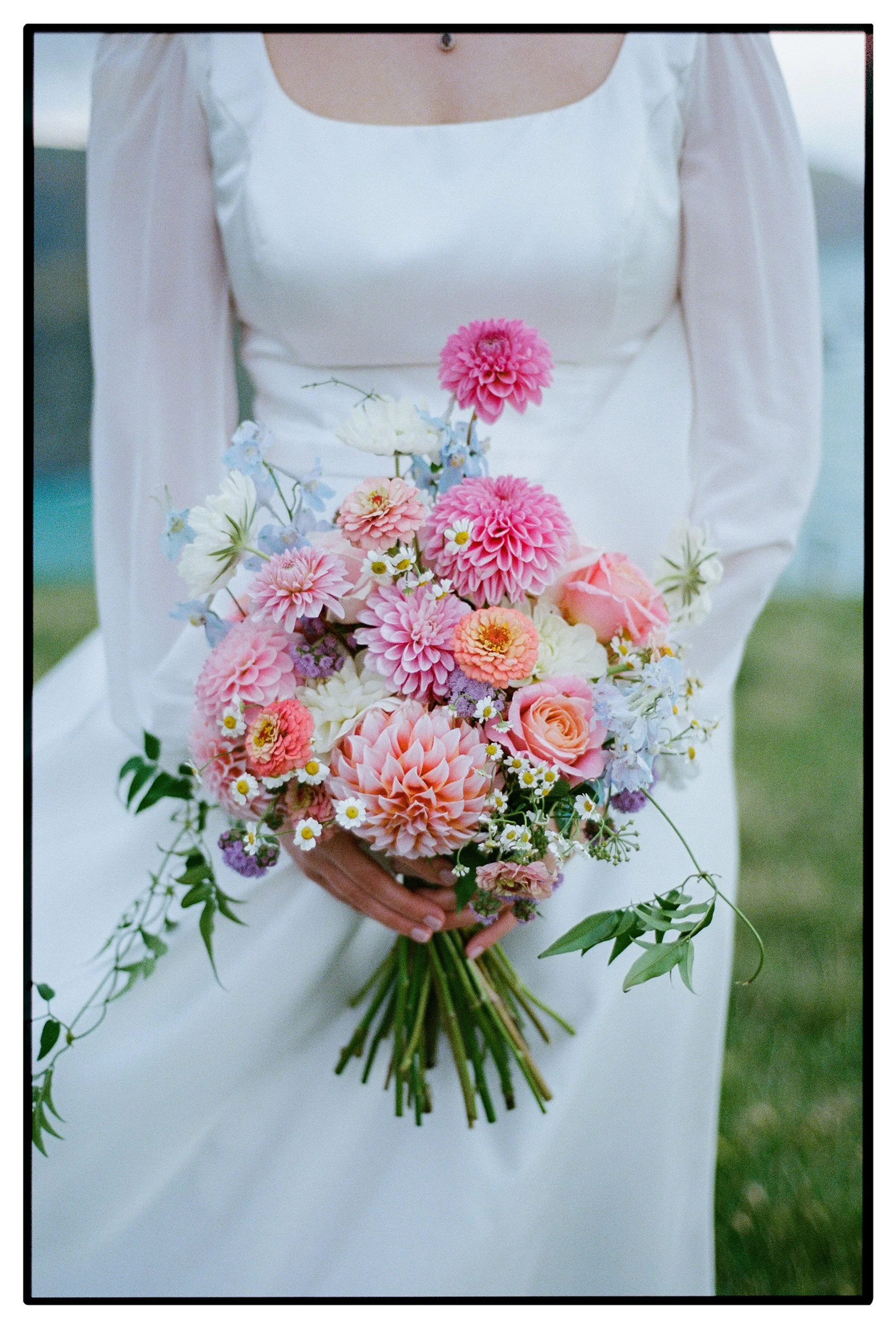 Film + Digital Wedding Photographer Christchurch - Accrington Farmhouse Wedding, Banks Peninsula, Christchurch, New Zealand. Film photo of bride’s white wedding dress and colourful bouquet with pink and orange flowers on a cliff by the ocean.