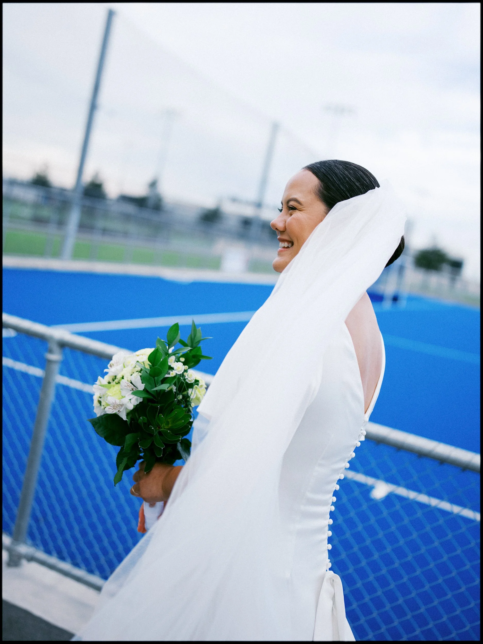 Film + Digital Wedding Photographer Christchurch - Backyard Wedding, Christchurch, Canterbury, New Zealand. Bride smiling and walking while holding bouquet of white flowers near blue hockey field. Editorial wedding photography.