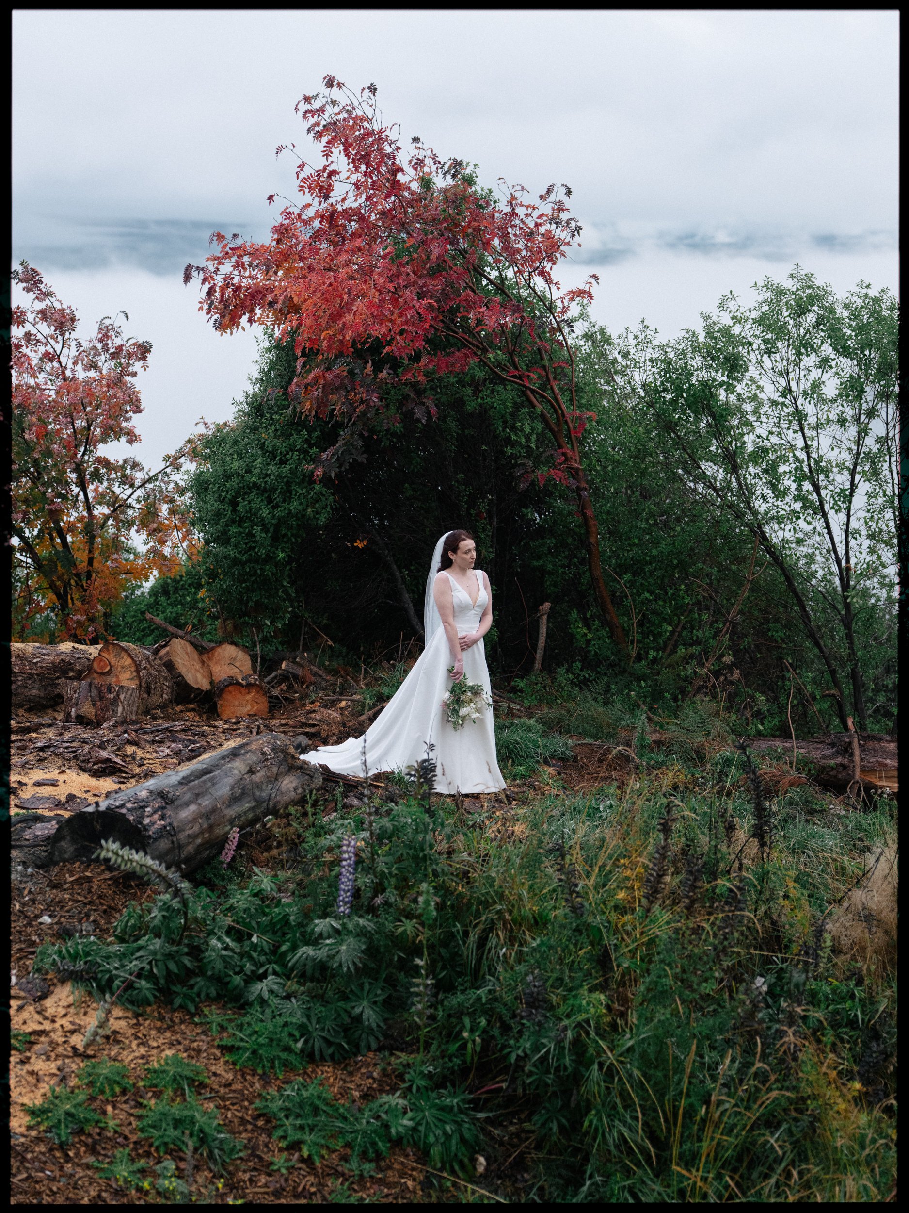 Film + Digital Wedding Photographer Queenstown - Kāmana Lakehouse, Queenstown, Otago, New Zealand. Bride standing in a wooded area with colourful autumn trees, holding a bouquet, wearing a white wedding gown and veil. Dramatic moody landscape.