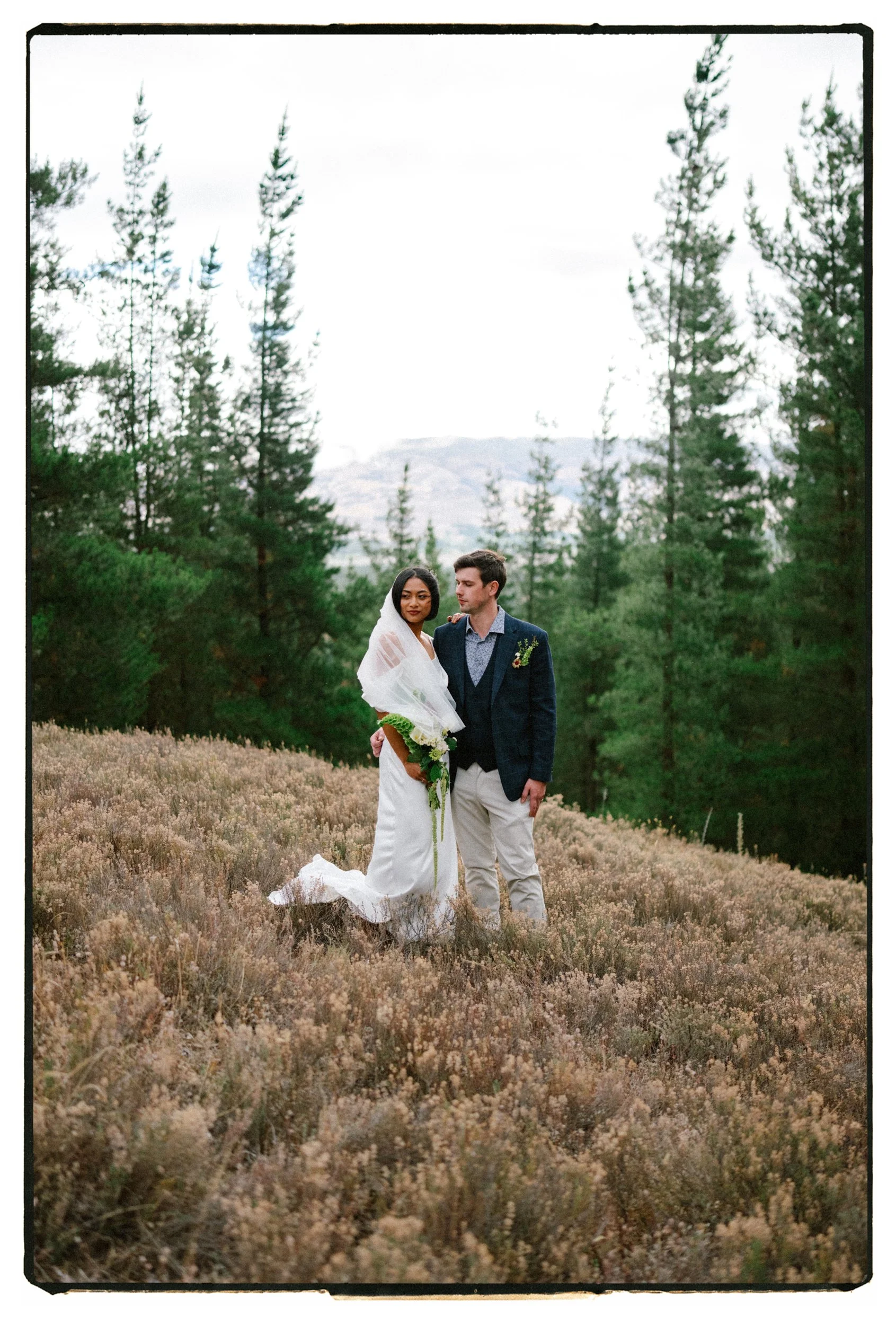 Film + Digital Wedding Photographer Christchurch - Alexandra Wedding, Otago, New Zealand. Moody photo of bride and groom standing in a grassy field with a backdrop of green pine trees and distant mountains on rainy day.