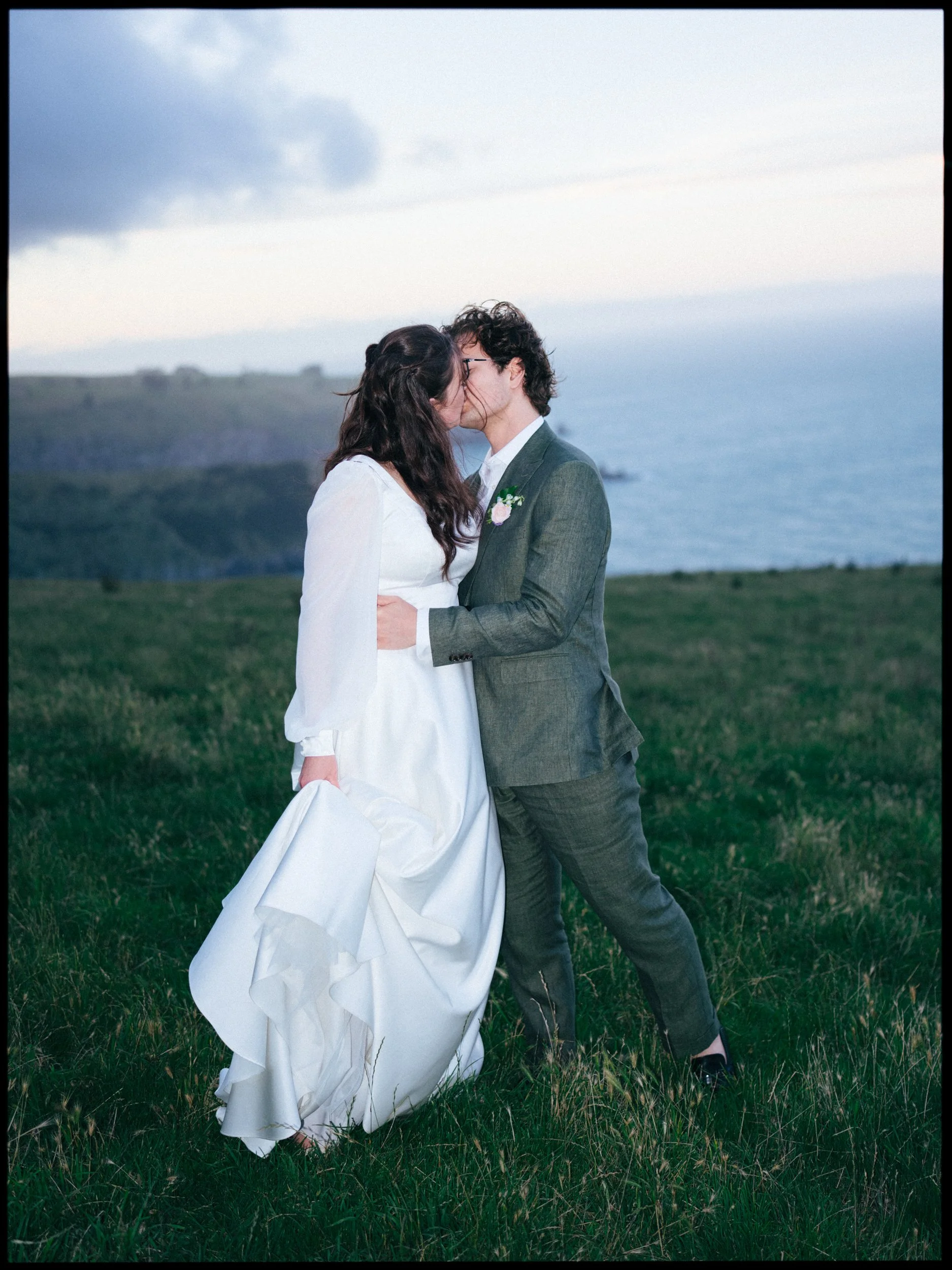 Accrington Farmhouse Wedding, Banks Peninsula, Christchurch, New Zealand. Bride and groom sharing a kiss outdoors on a grassy field with the ocean and cliffs in the background.