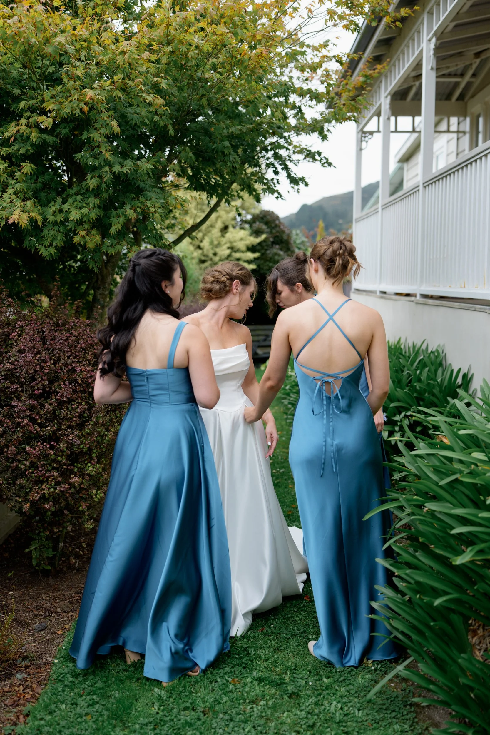 Film + Digital Wedding Photographer Dunedin - Lochend Woolshed, Dunedin, Otago, New Zealand. Bride and bridesmaids standing outdoors on a garden path, surrounded by greenery and trees near a white building.