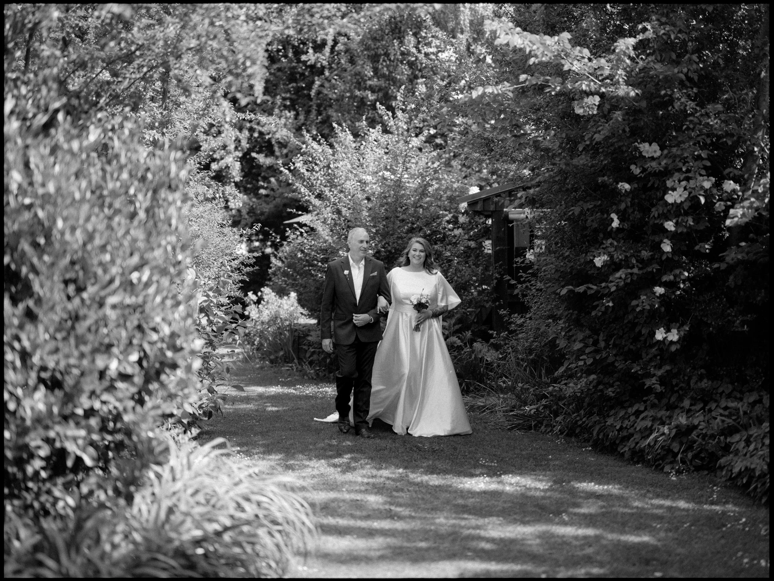 Kaiapoi Wedding, Canterbury, New Zealand. A black and white photo of a bride walking down the aisle arm-in-arm with her father on a garden pathway surrounded by lush greenery and flowering bushes.