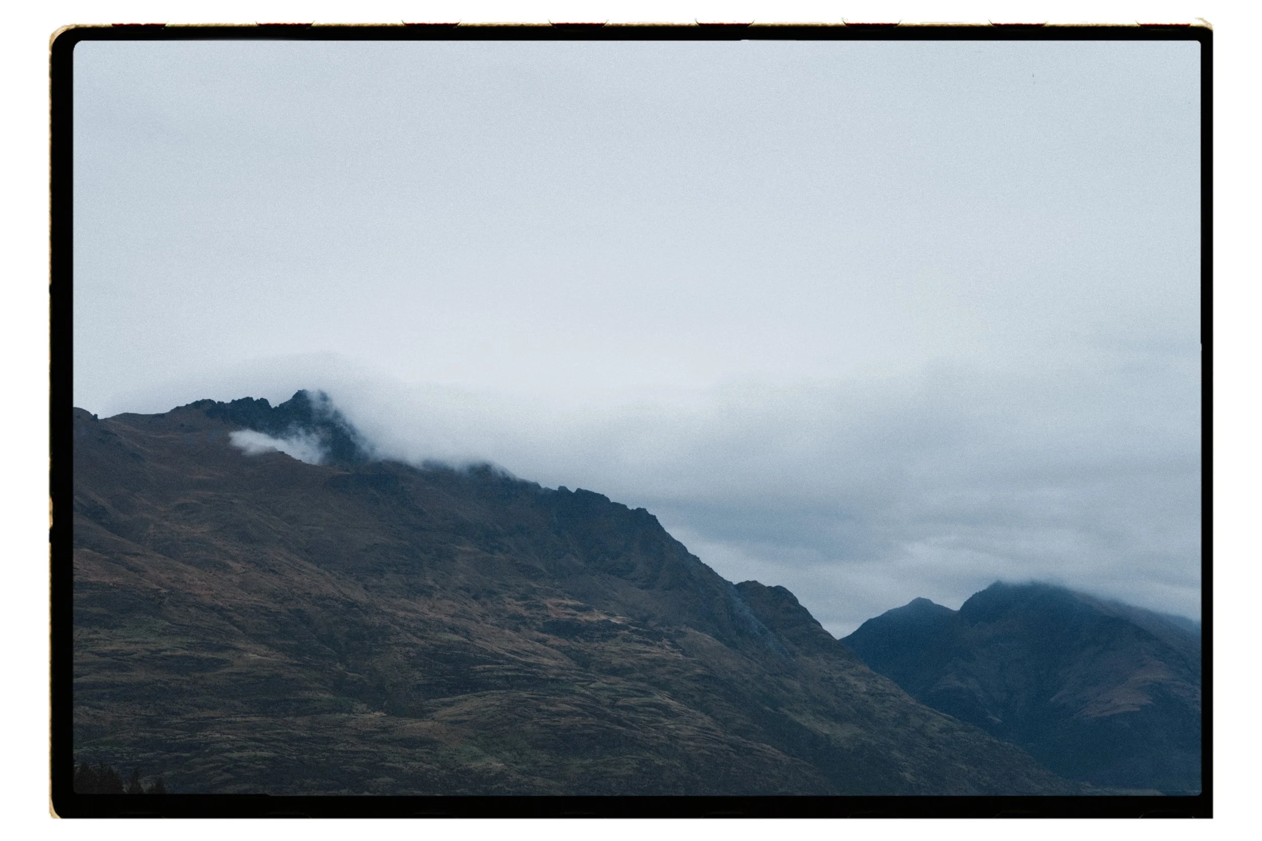 Film + Digital Wedding Photographer Queenstown - Kāmana Lakehouse, Queenstown, Otago, New Zealand. Mountain landscape with rugged peaks, some shrouded in clouds, under an overcast sky surrounding Moke Lake.