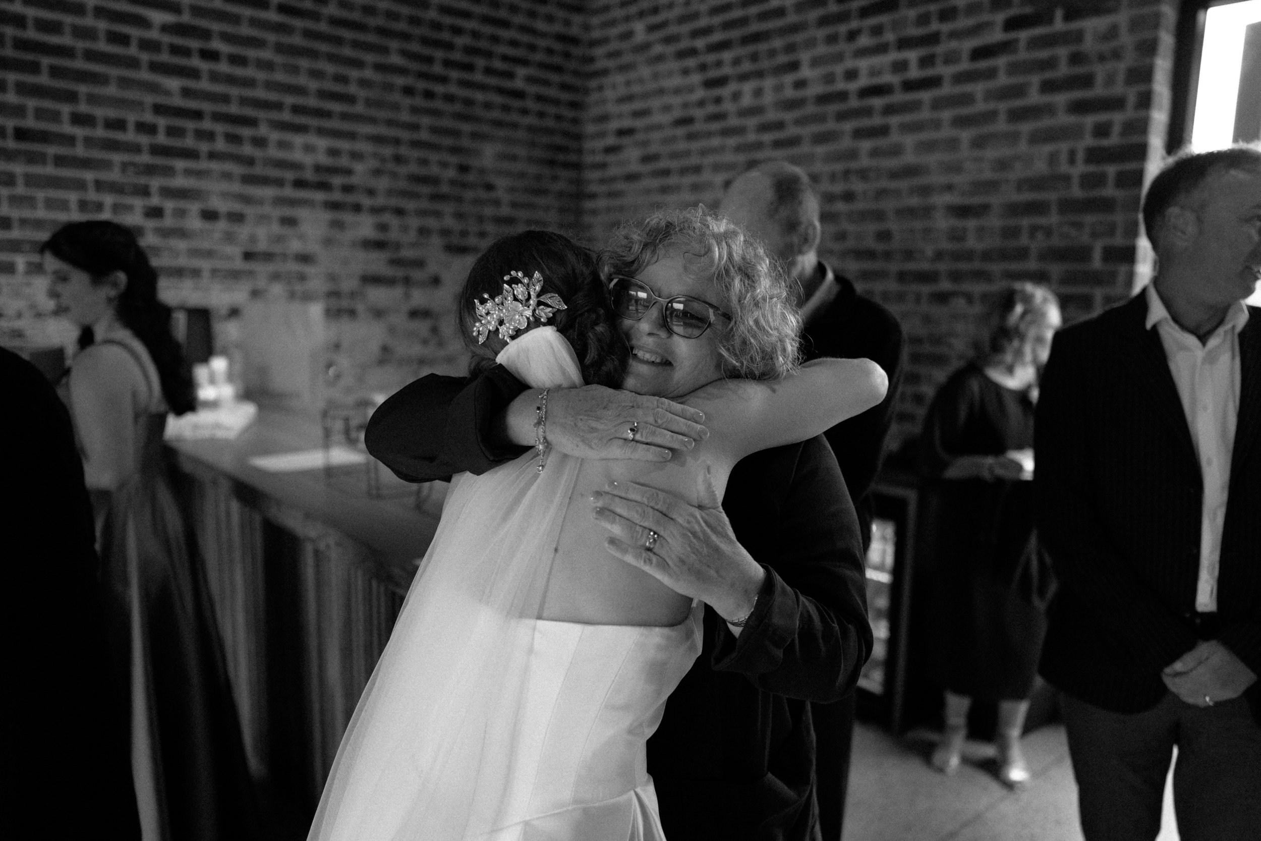 Film + Digital Wedding Photographer Dunedin - Lochend Woolshed, Dunedin, Otago, New Zealand. Candid emotional photo of elderly woman embracing bride at wedding reception, with other guests in the background near a brick wall.
