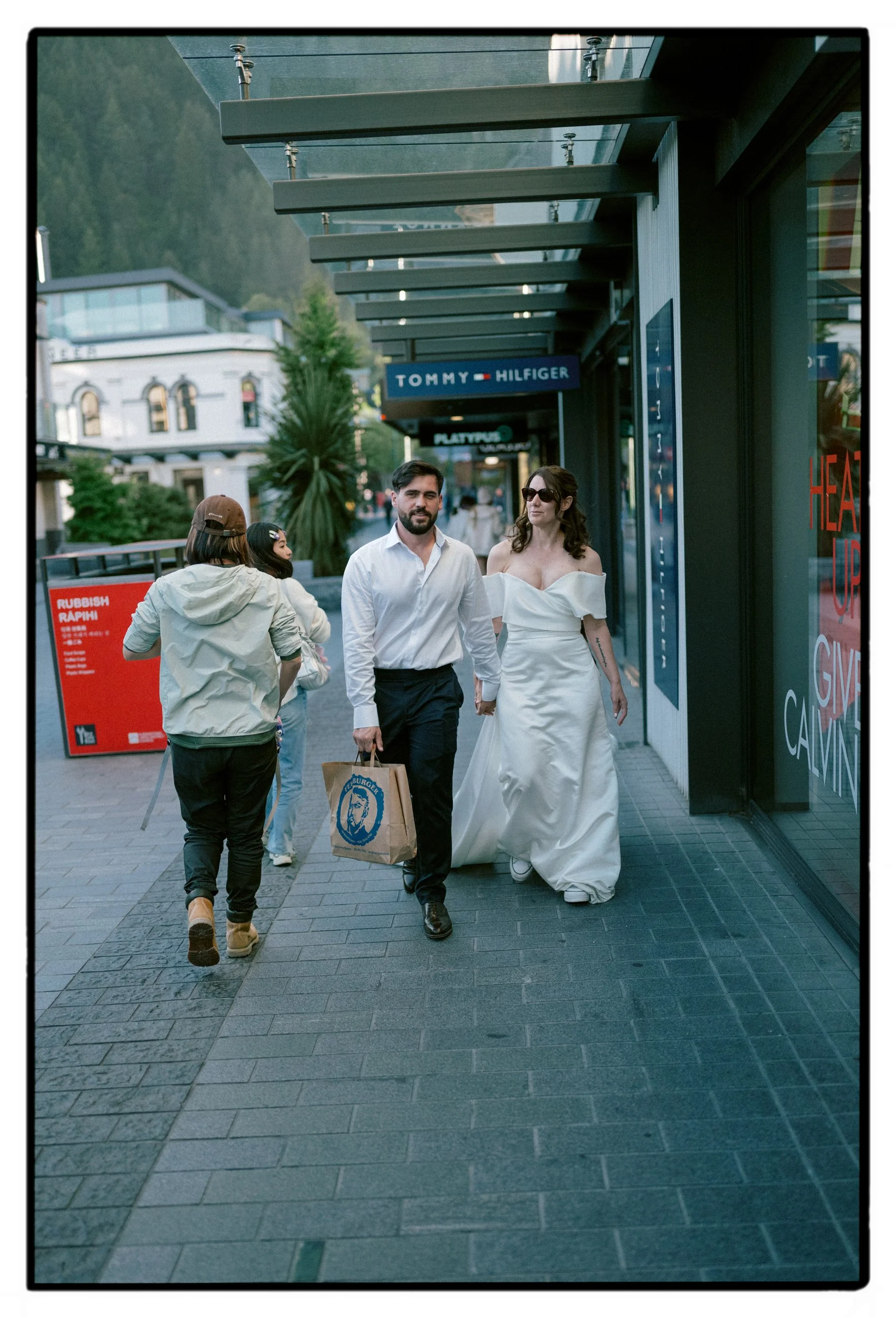 Film + Digital Wedding Photographer Christchurch - Queenstown Elopement, Otago, New Zealand. Bride and groom walking down street holding hands and carrying Fergburger bag