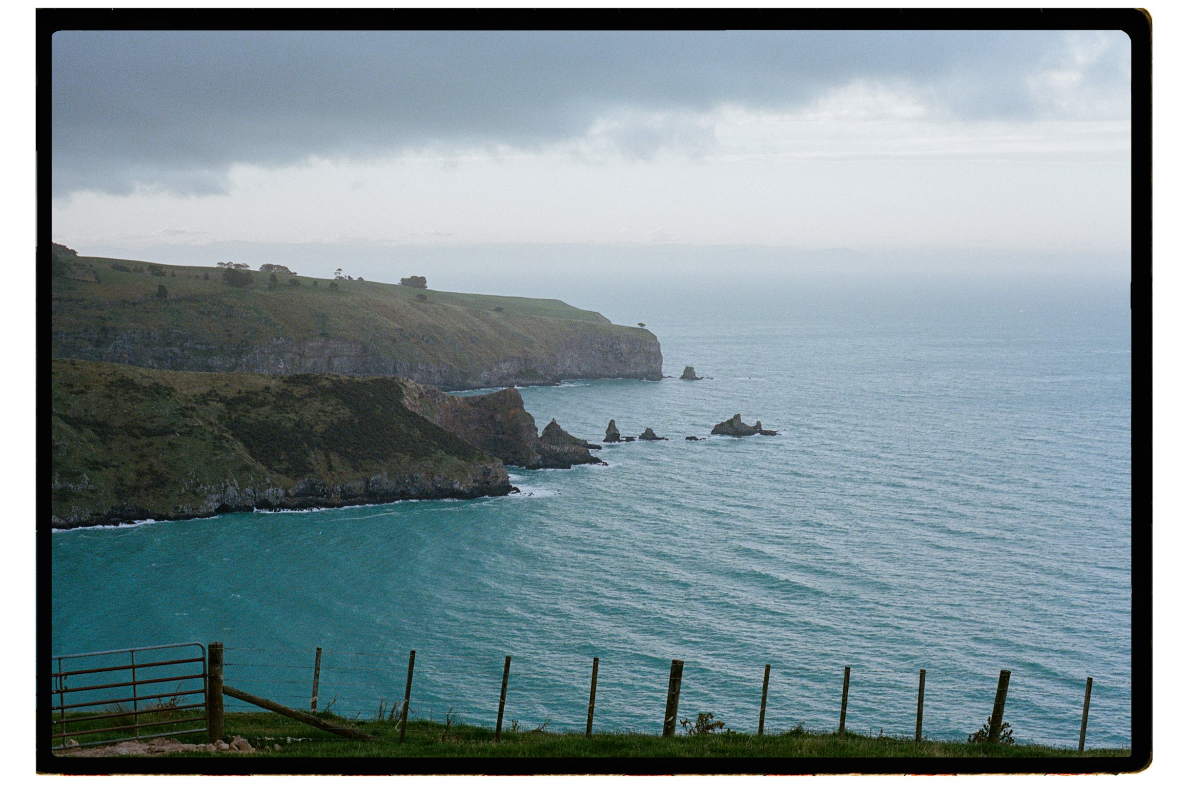Accrington Farmhouse Wedding, Banks Peninsula, Christchurch, New Zealand. A rugged coastline with green hills and cliffs dropping into the ocean, with rocks jutting out into the water under a cloudy sky.
