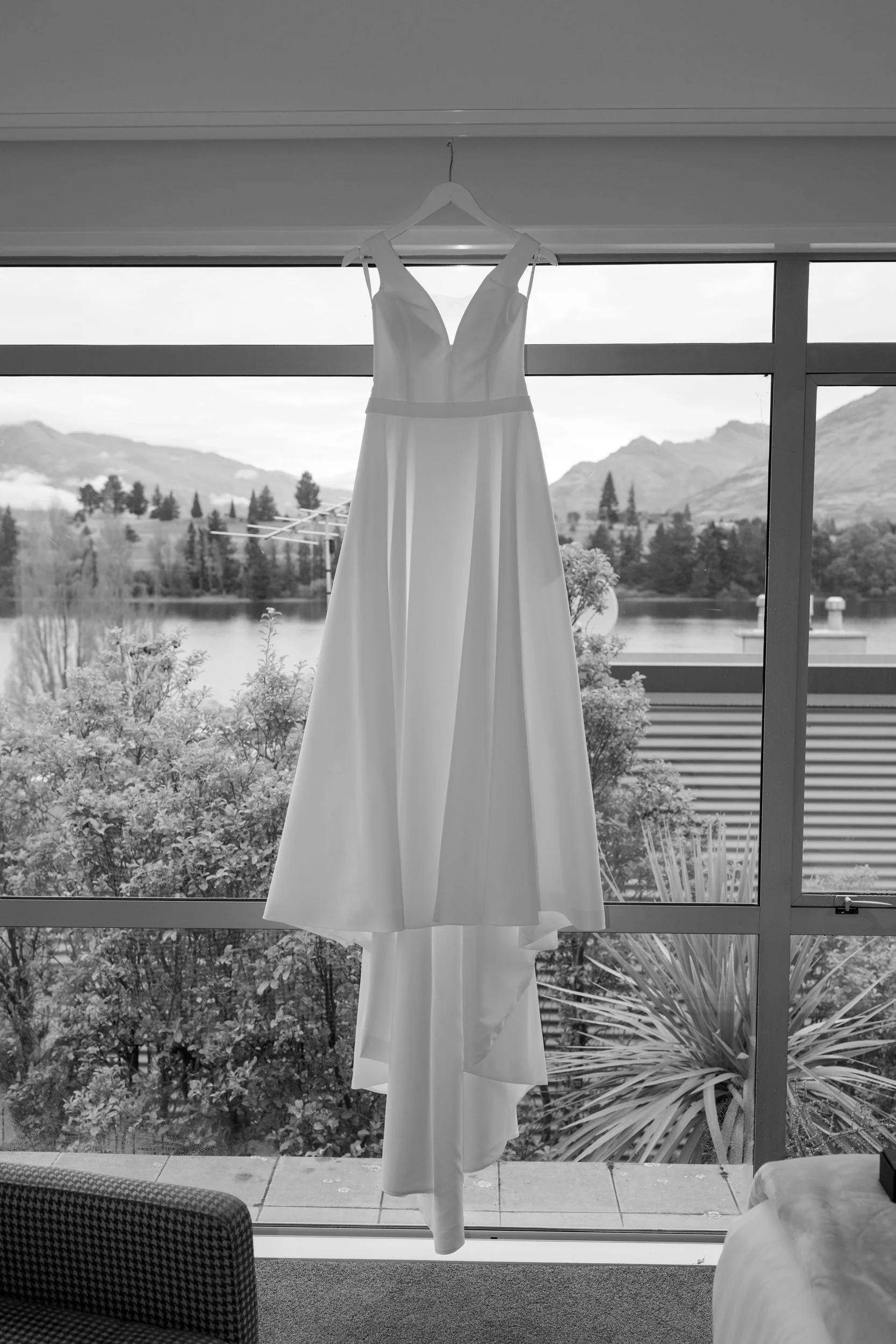 Film + Digital Wedding Photographer Queenstown - Kāmana Lakehouse, Queenstown, Otago, New Zealand. Wedding dress hanging on a hanger in front of a large window with a view of the mountains and Lake Wakatipu.