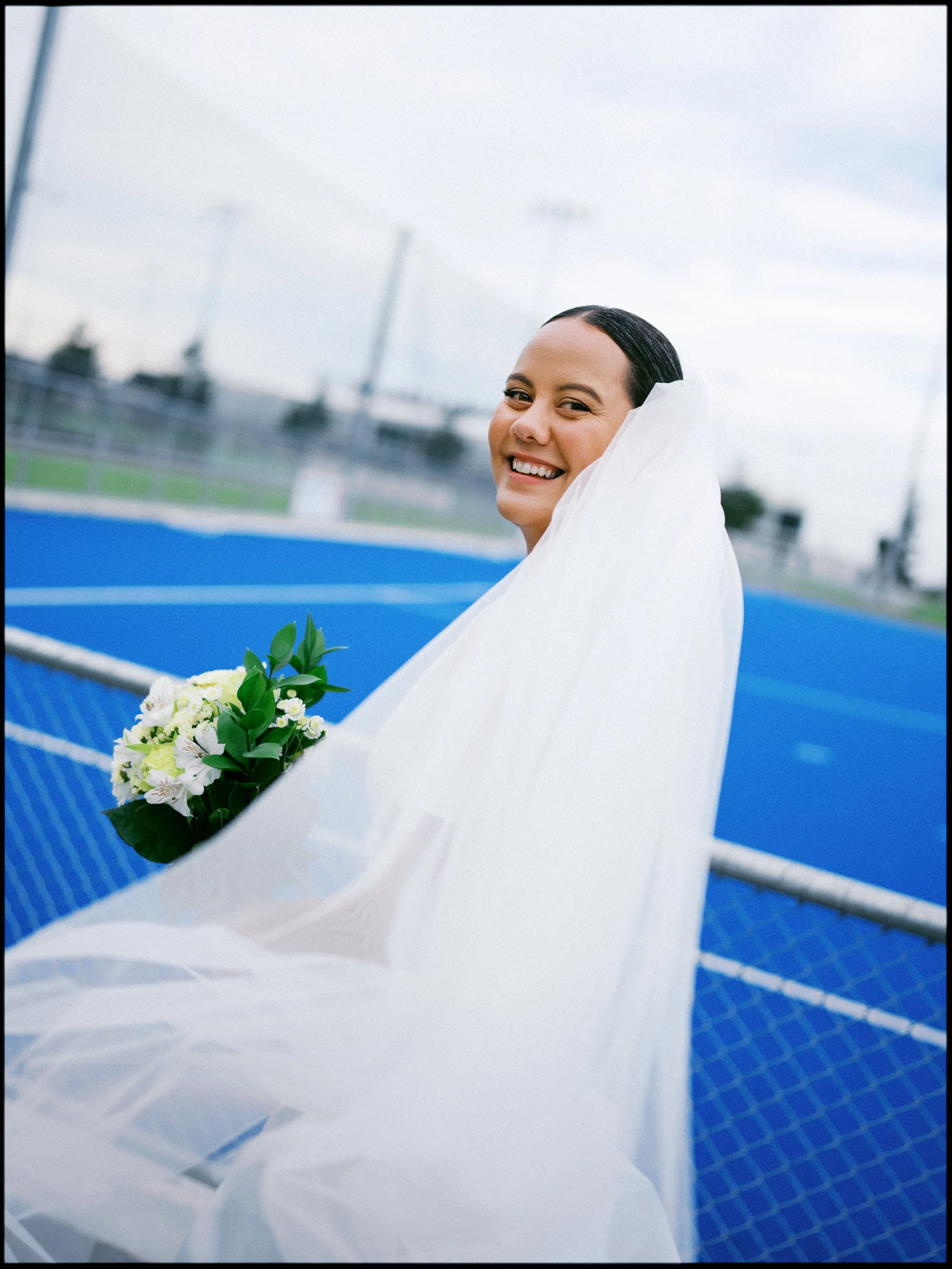 Film + Digital Wedding Photographer Christchurch - Backyard Wedding, Christchurch, Canterbury, New Zealand. Bride smiling looking at camera holding bouquet of white flowers near blue hockey field. Editorial wedding photography.