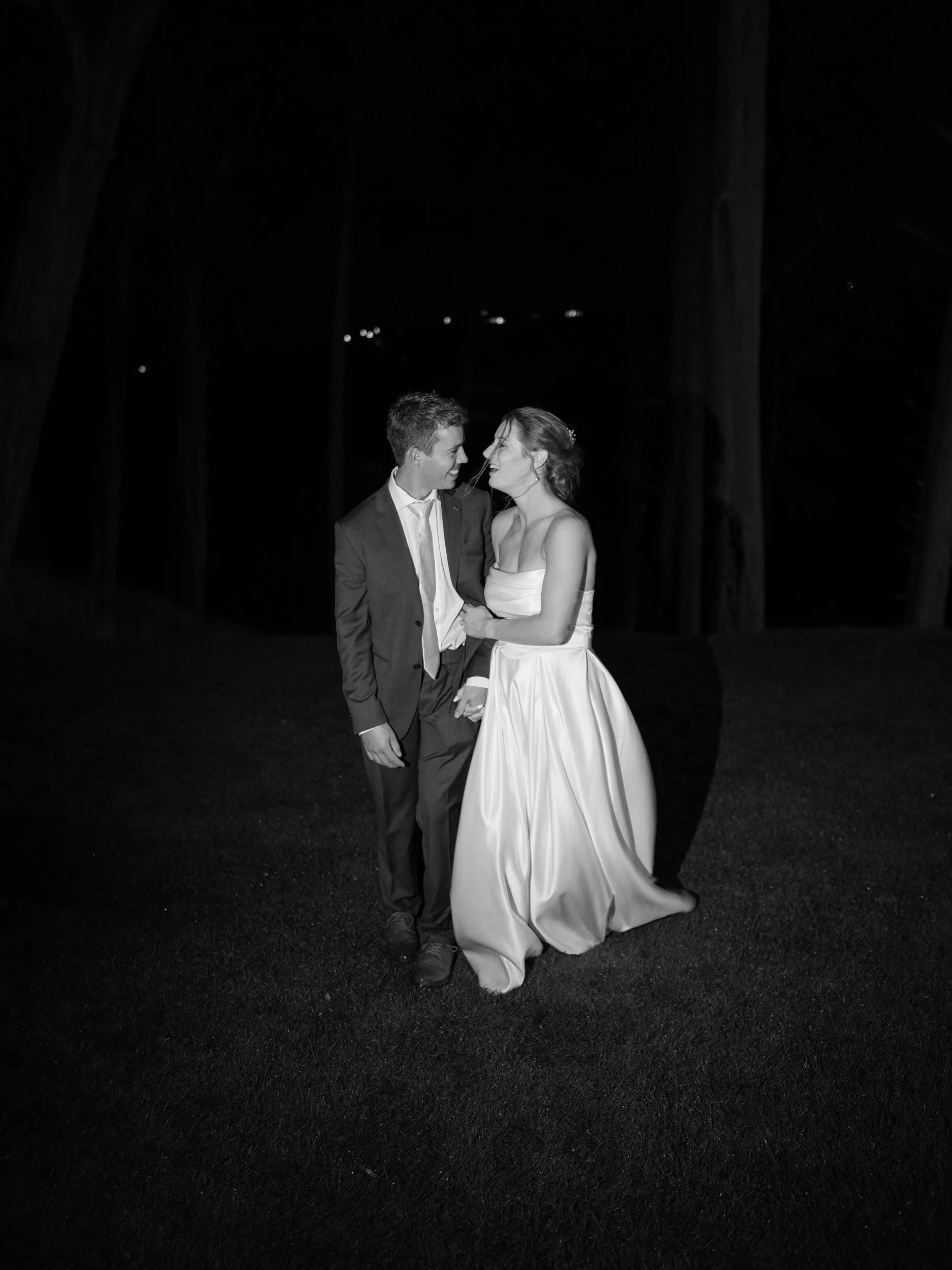 Film + Digital Wedding Photographer Dunedin - Lochend Woolshed, Dunedin, Otago, New Zealand. A black-and-white flash photo of bride and groom at night outdoors among tall trees.