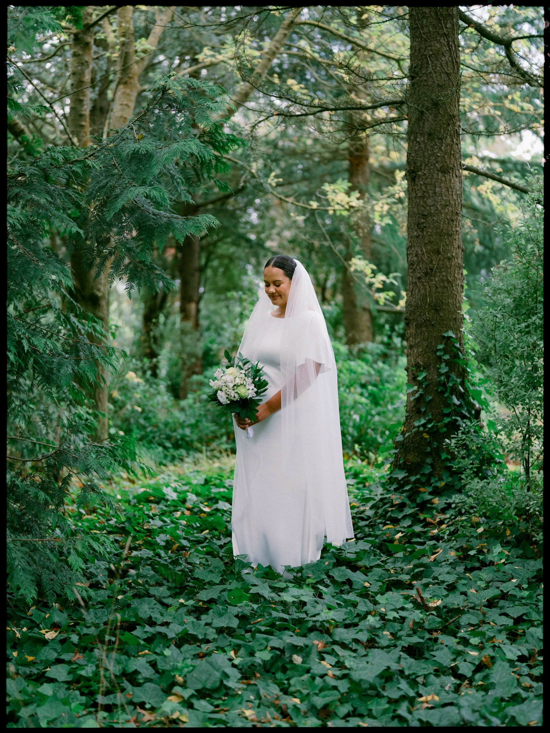 Film + Digital Wedding Photographer Christchurch - Backyard Wedding, Christchurch, Canterbury, New Zealand. Bride in white wedding gown and veil holding a bouquet of white flowers, standing in a lush green forest surrounded by trees and ivy.