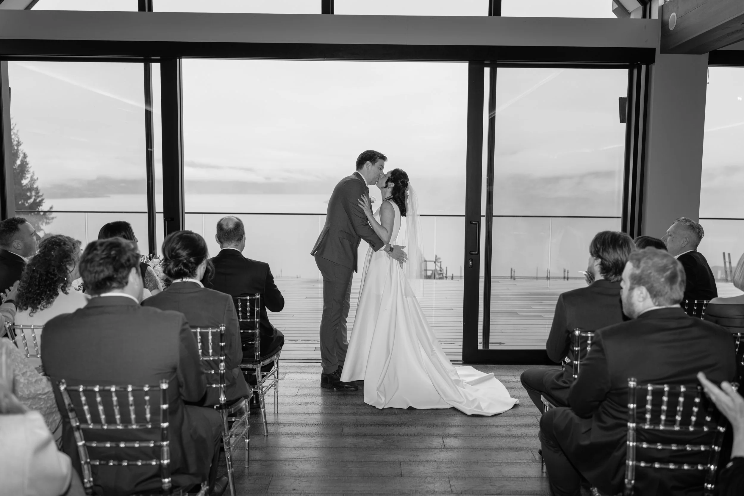 Film + Digital Wedding Photographer Queenstown - Kāmana Lakehouse, Queenstown, Otago, New Zealand. Black and white photograph of indoor wedding ceremony overlooking Lake Wakatipu. The bride and groom are kissing, surrounded by seated guests.