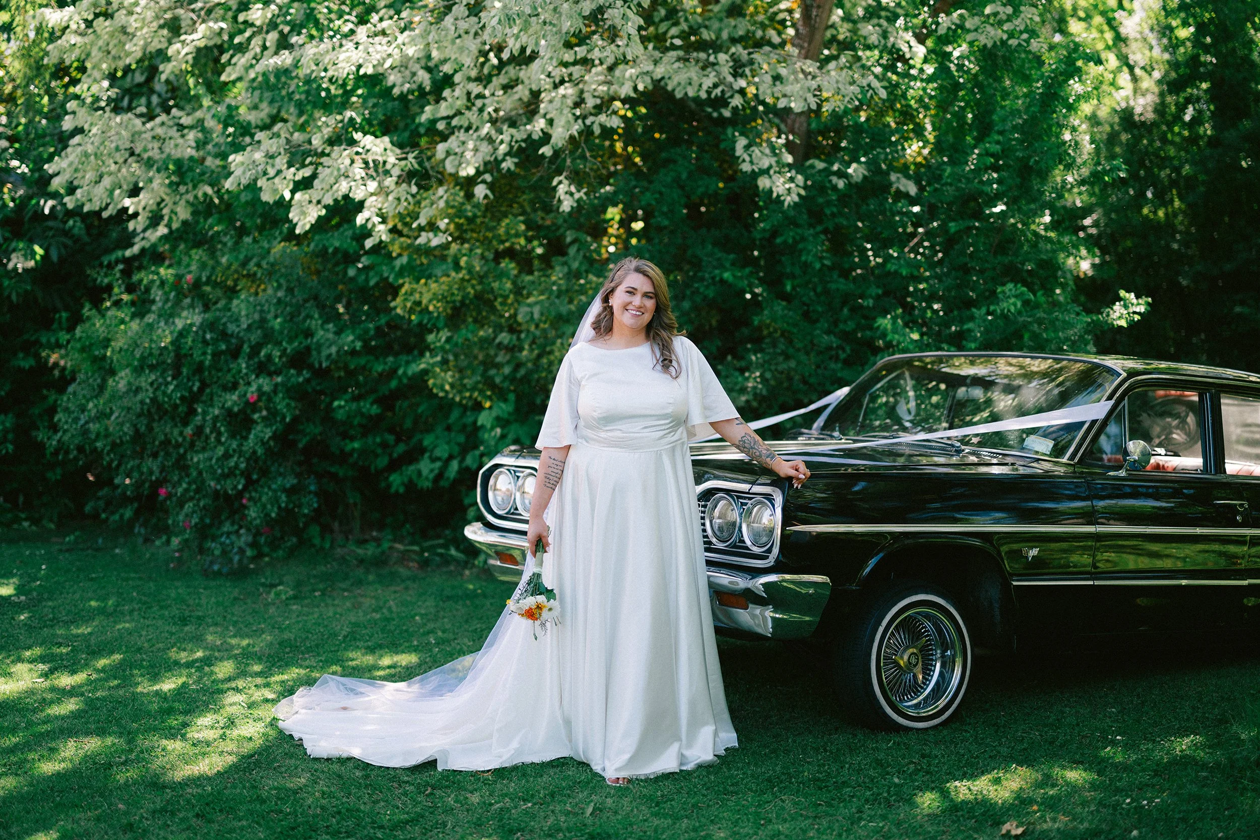 Film + Digital Wedding Photographer Christchurch - Kaiapoi Wedding, Canterbury, New Zealand. Portrait of bride in front of a black 1964 Chevrolet Impala in a lush green garden.