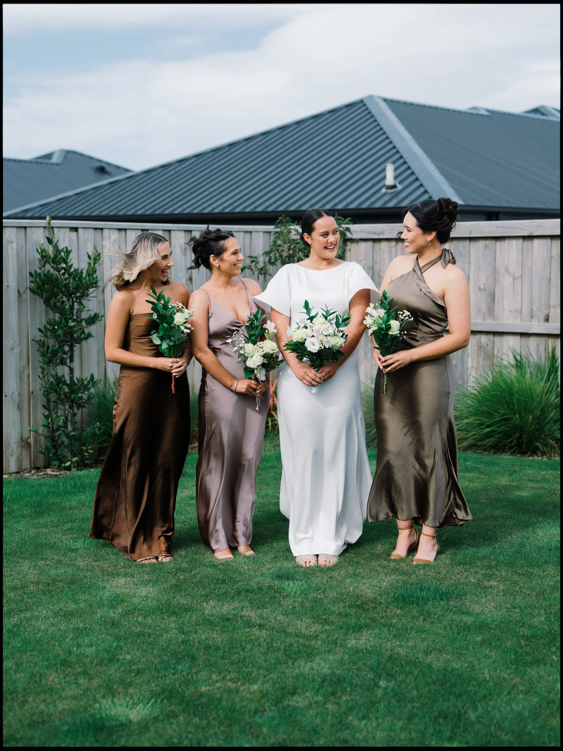 Film + Digital Wedding Photographer Christchurch - Backyard Wedding, Christchurch, Canterbury, New Zealand. Bride and bridesmaids holding bouquets, standing on backyard green lawn in front of wooden fence smiling and talking to each other.