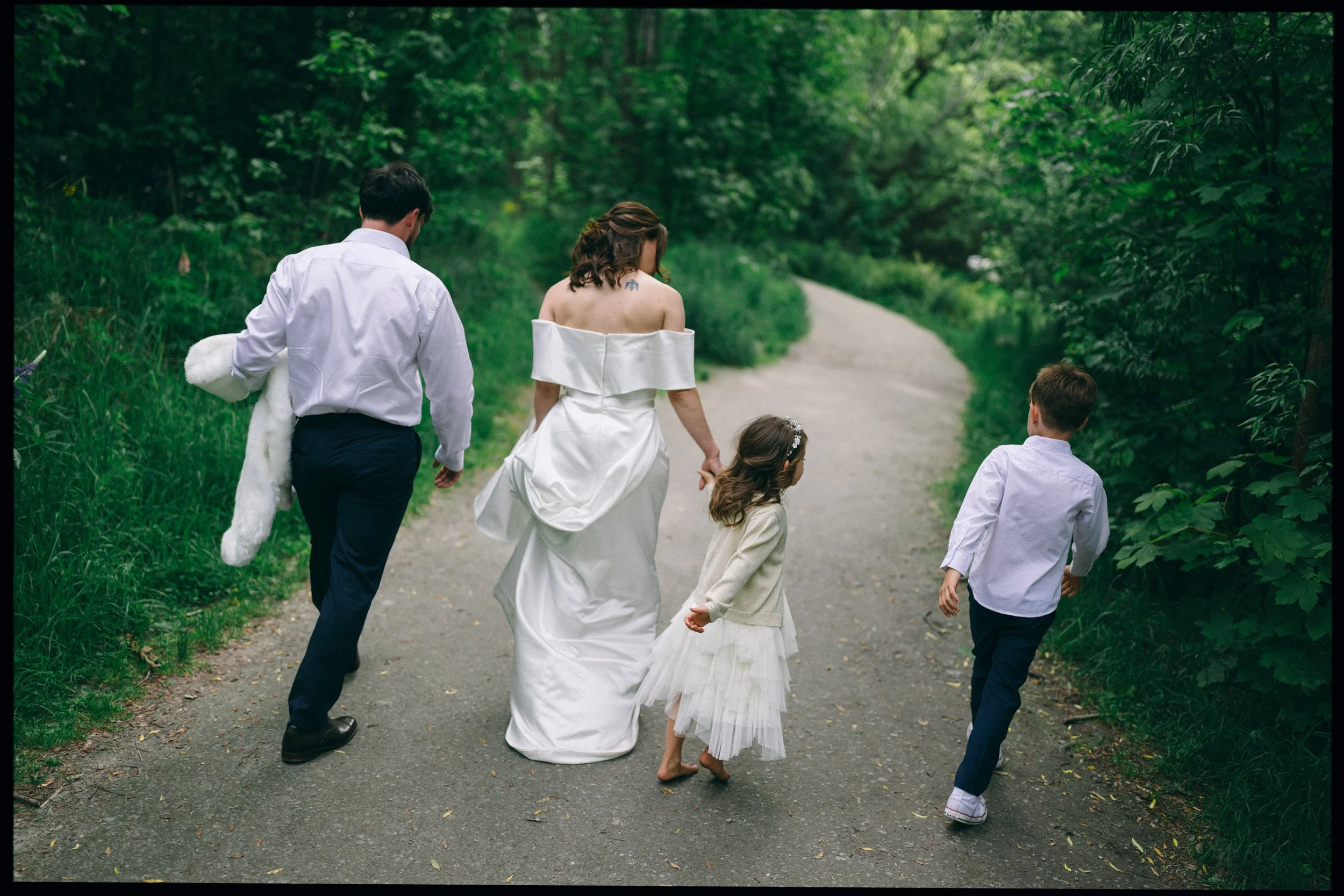 Queenstown Elopement, Otago, New Zealand. Bride and groom walking outdoors with their two children, a boy and a girl, on a forested path along the Arrow river in Arrowtown.