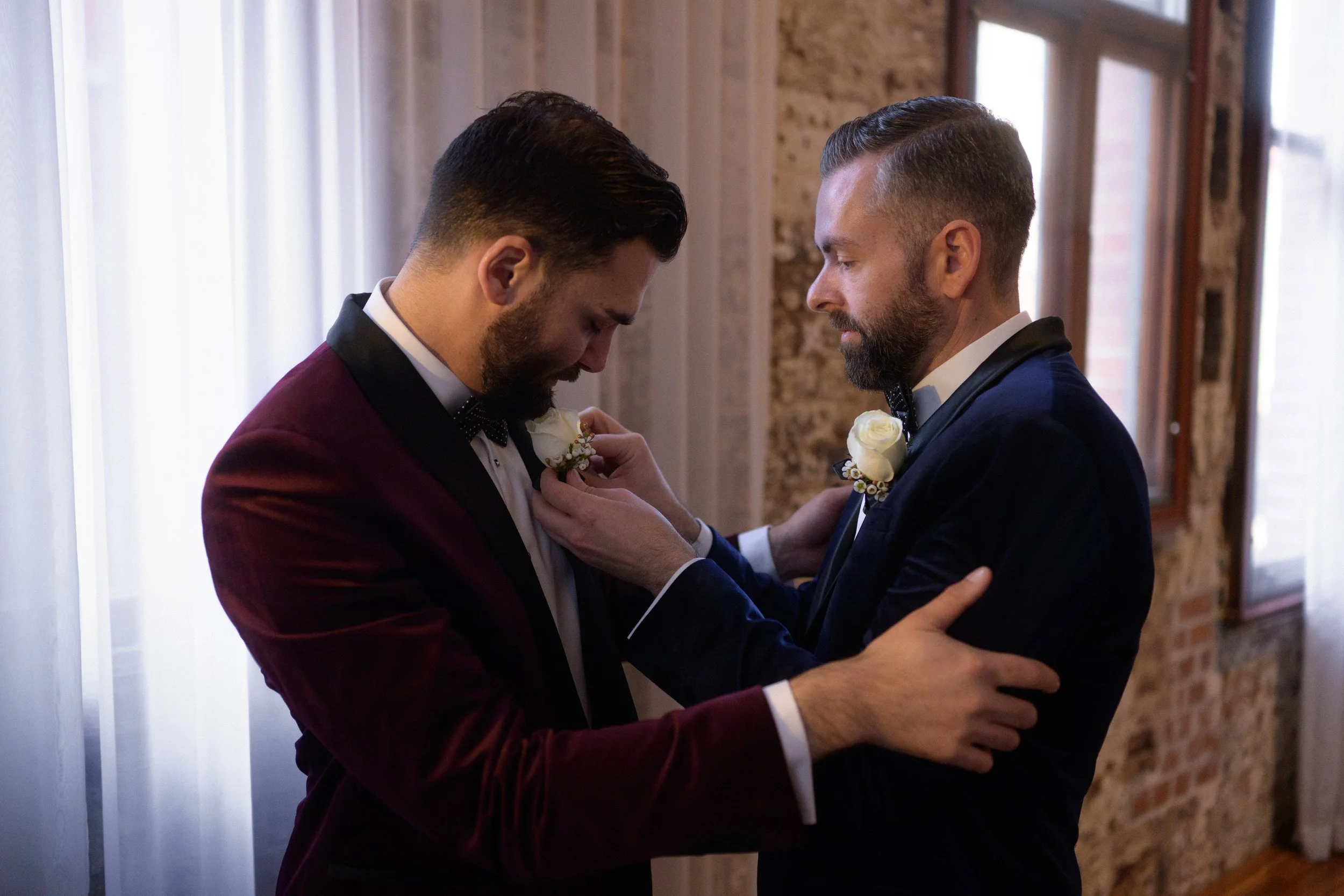 Grooms helping each other get ready for their wedding. One is wearing a burgundy suit and the other a blue one.