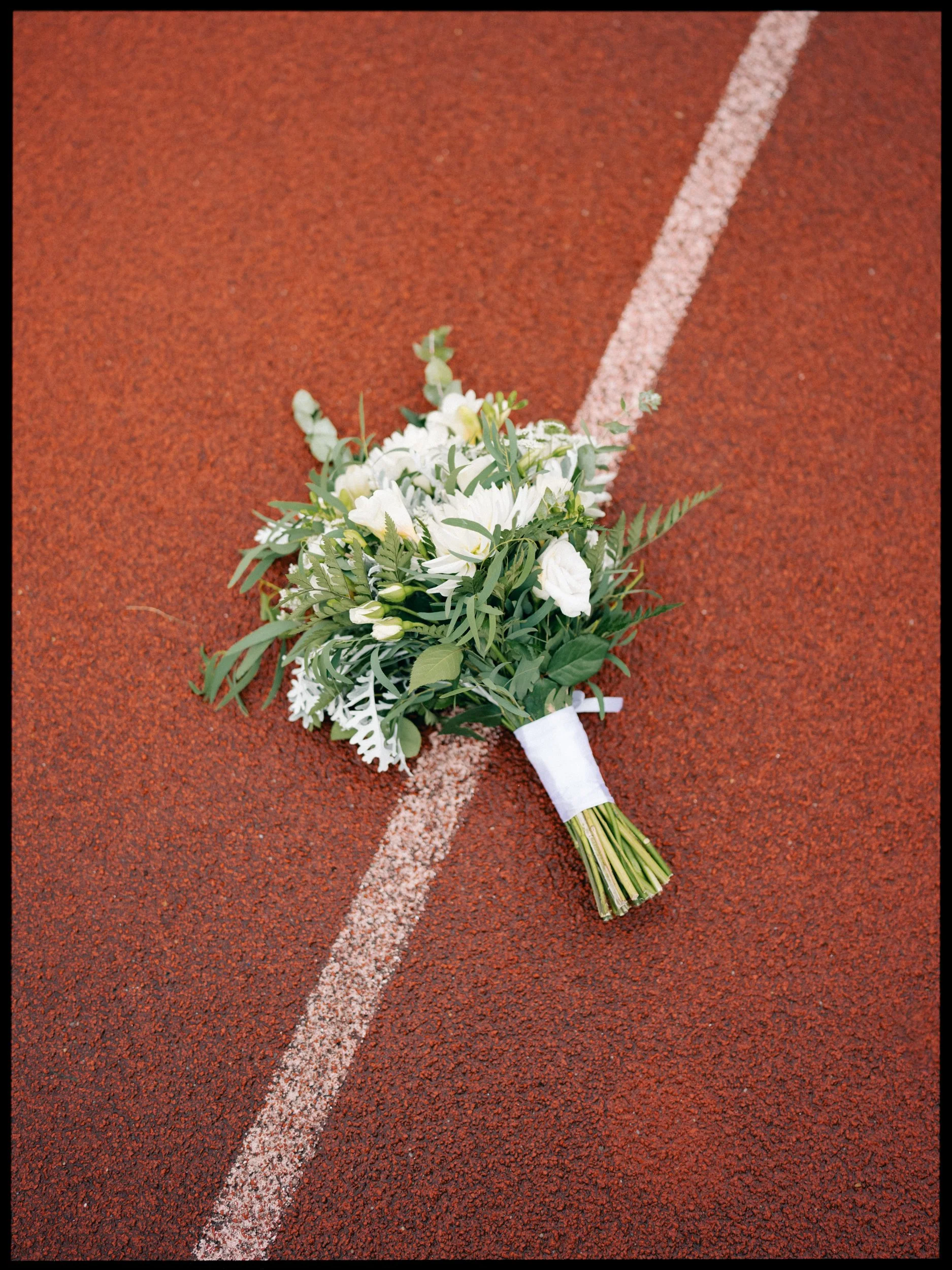 Film + Digital Wedding Photographer Dunedin - Lochend Woolshed, Dunedin, Otago, New Zealand. A bouquet of white flowers placed on a red running track at the Caledonian Ground. Editorial wedding photography.