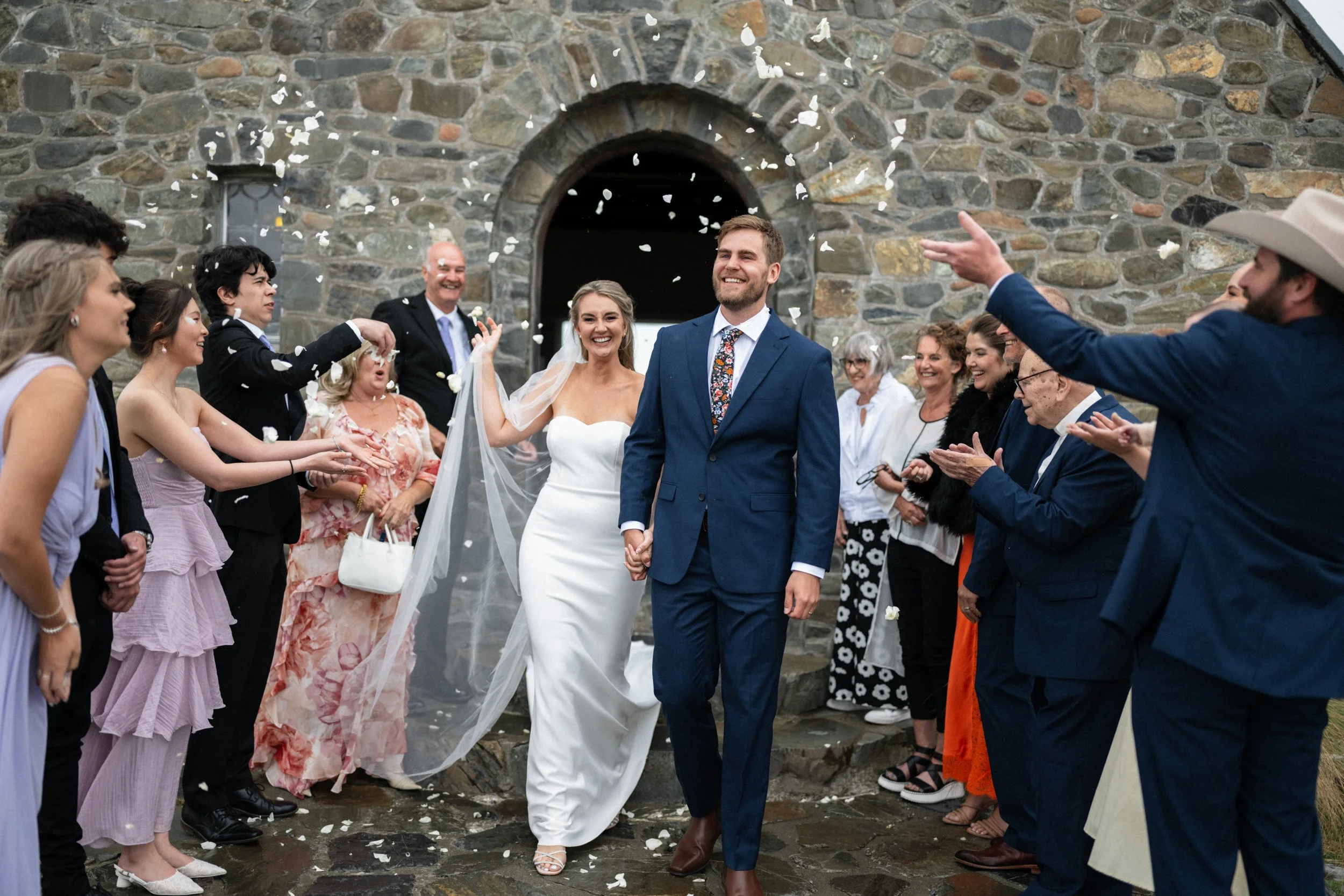 Film + Digital Wedding Photographer Christchurch - Lake Tekapo Wedding, Canterbury, New Zealand. Bride and groom smiling and walking out of the Church of the Good Shepherd on a rainy day, celebrated by wedding guests throwing white flower petals.
