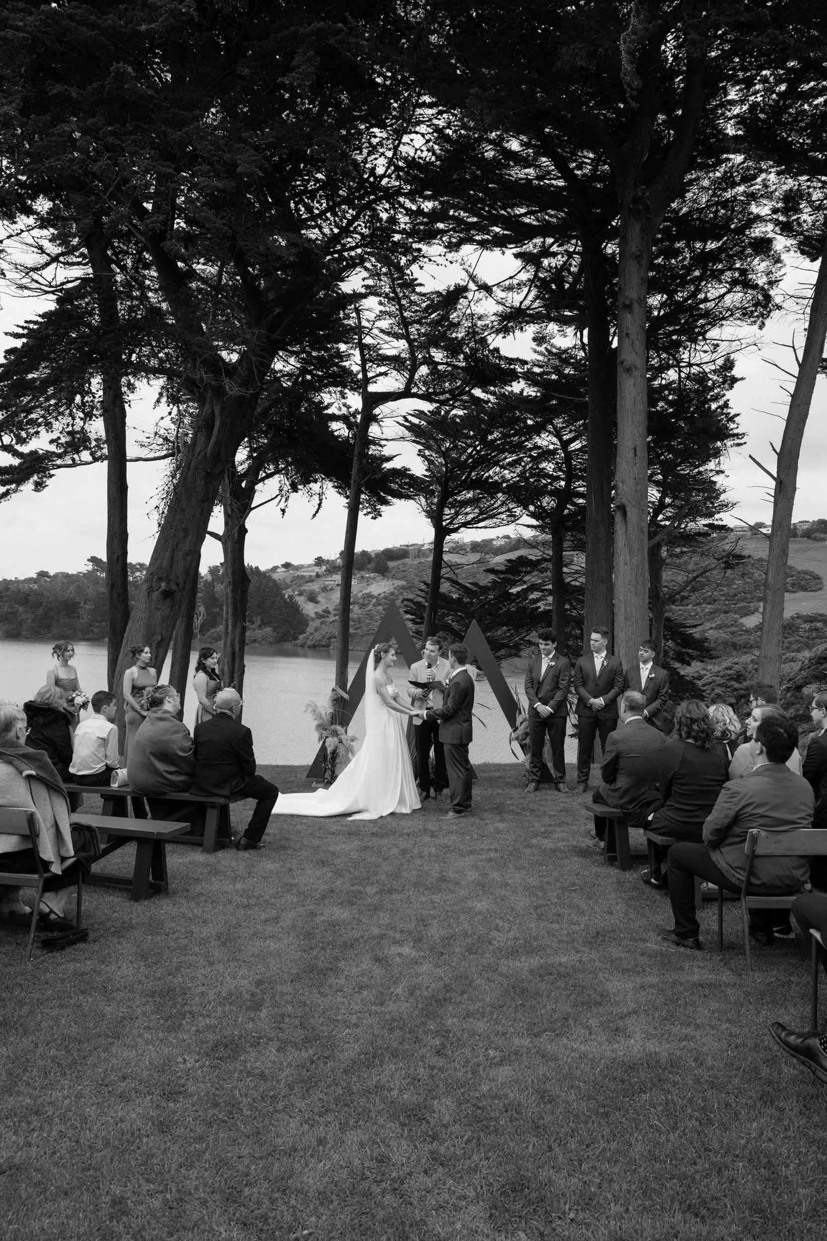 Film + Digital Wedding Photographer Dunedin - Lochend Woolshed, Dunedin, Otago, New Zealand. Wedding ceremony outdoors near lake, with tall trees in the background. Bride and groom are standing in front of celebrant with guests around them.