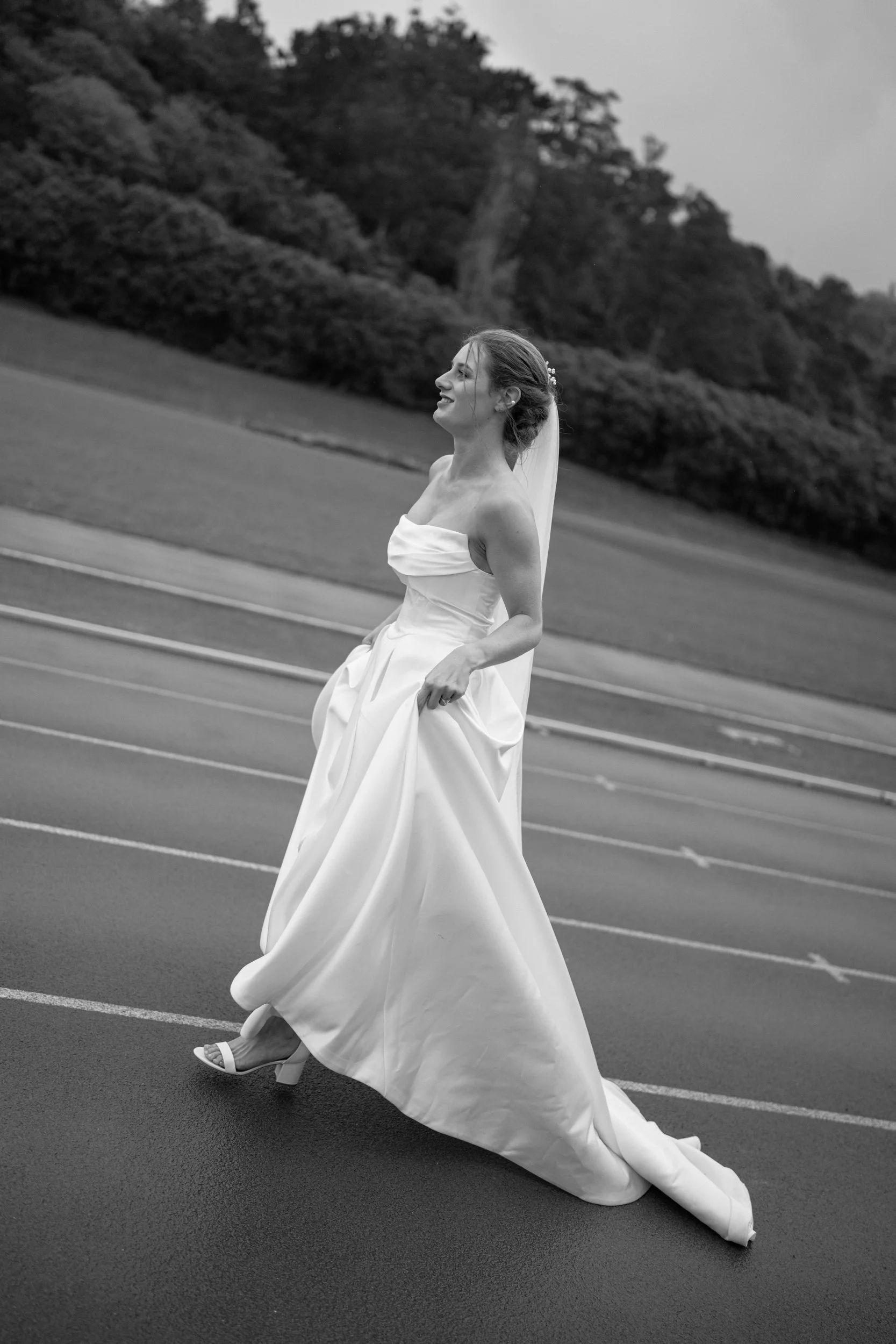 Film + Digital Wedding Photographer Dunedin - Lochend Woolshed, Dunedin, Otago, New Zealand. Black and white photo of bride in strapless wedding gown running on race track at Caledonian Ground. Editorial wedding photography.