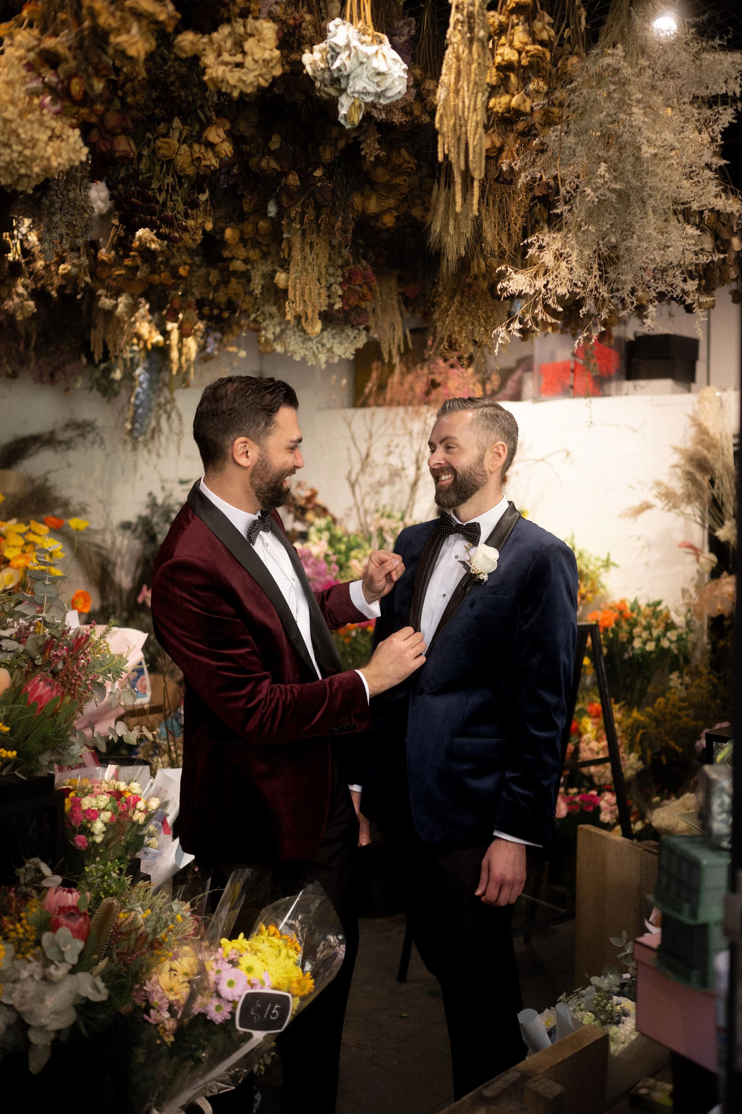 Grooms smiling at each other in colourful flower shop.