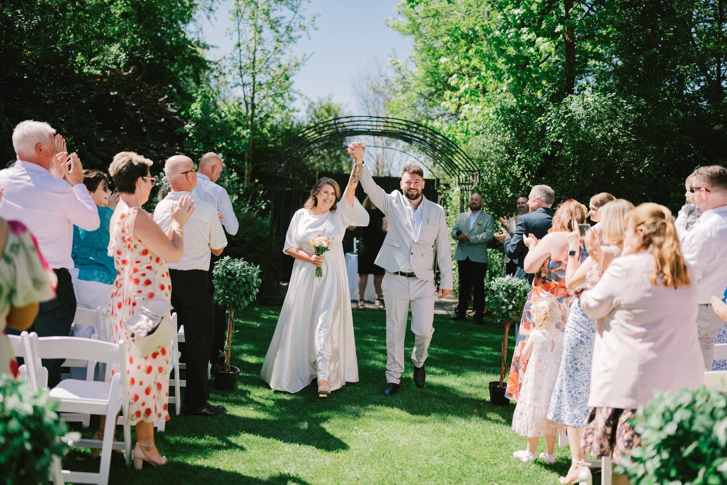 Kaiapoi Wedding, Canterbury, New Zealand.  A newlywed couple walks down the aisle, holding hands and raising intertwined arms in celebration, surrounded by guests clapping and celebrating outdoors on a sunny day.
