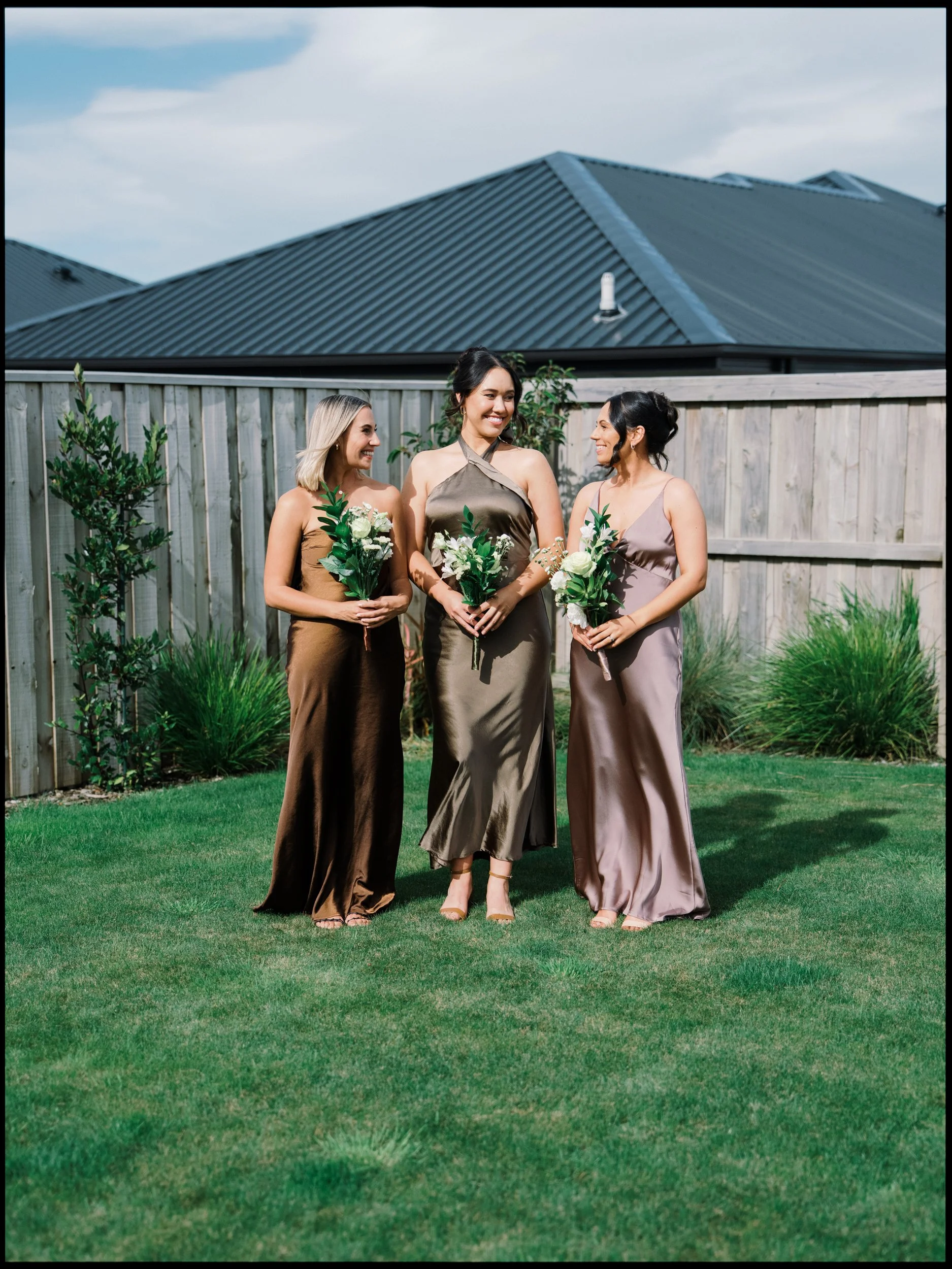 Film + Digital Wedding Photographer Christchurch - Backyard Wedding, Christchurch, Canterbury, New Zealand. Three bridesmaids holding bouquets, standing on a lawn in front of a wooden fence.