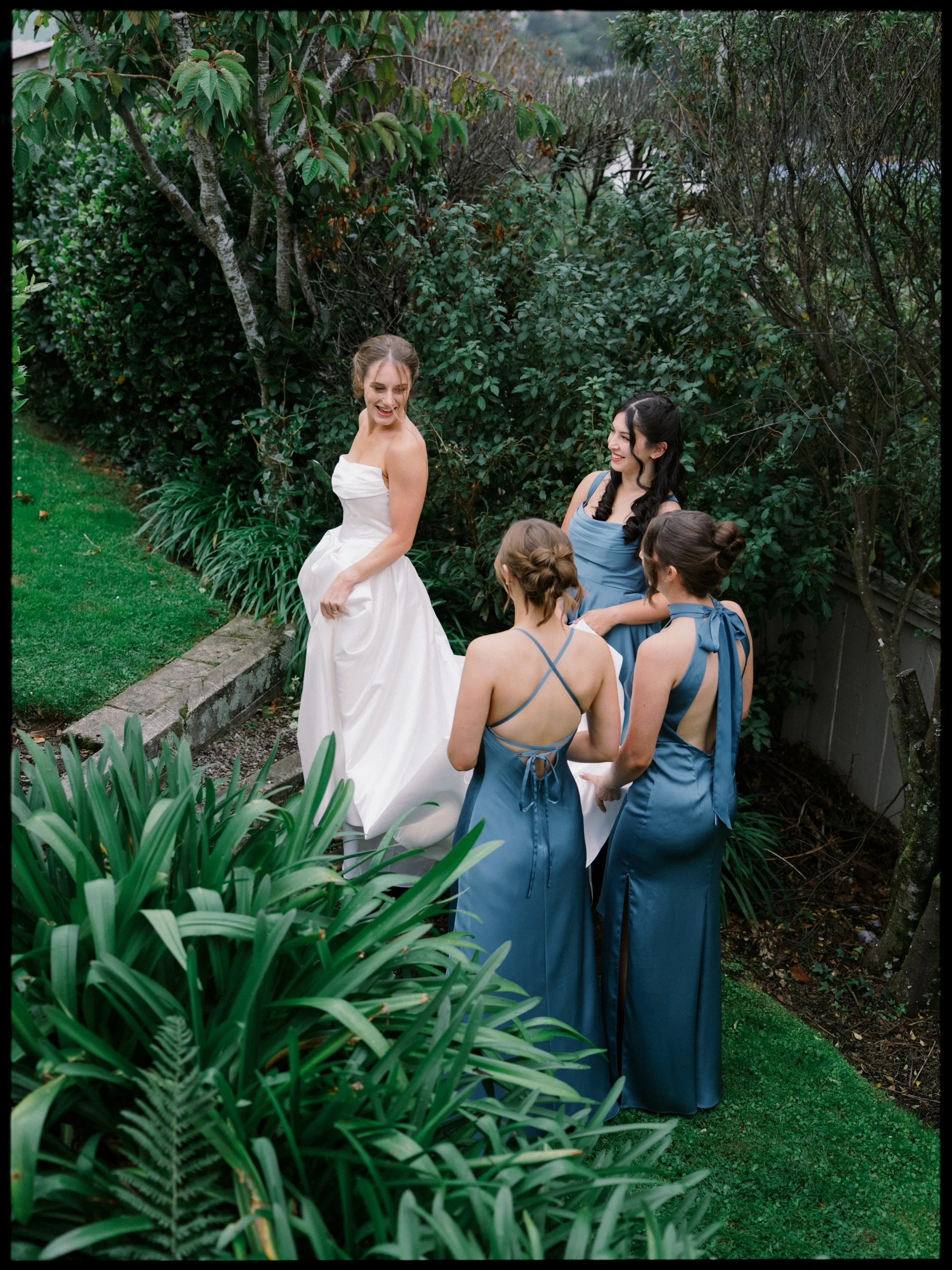 Film + Digital Wedding Photographer Dunedin - Lochend Woolshed, Dunedin, Otago, New Zealand. Bride in white strapless wedding dress laughing and looking at bridesmaids in matching blue dresses standing in a lush garden with green bushes and trees.