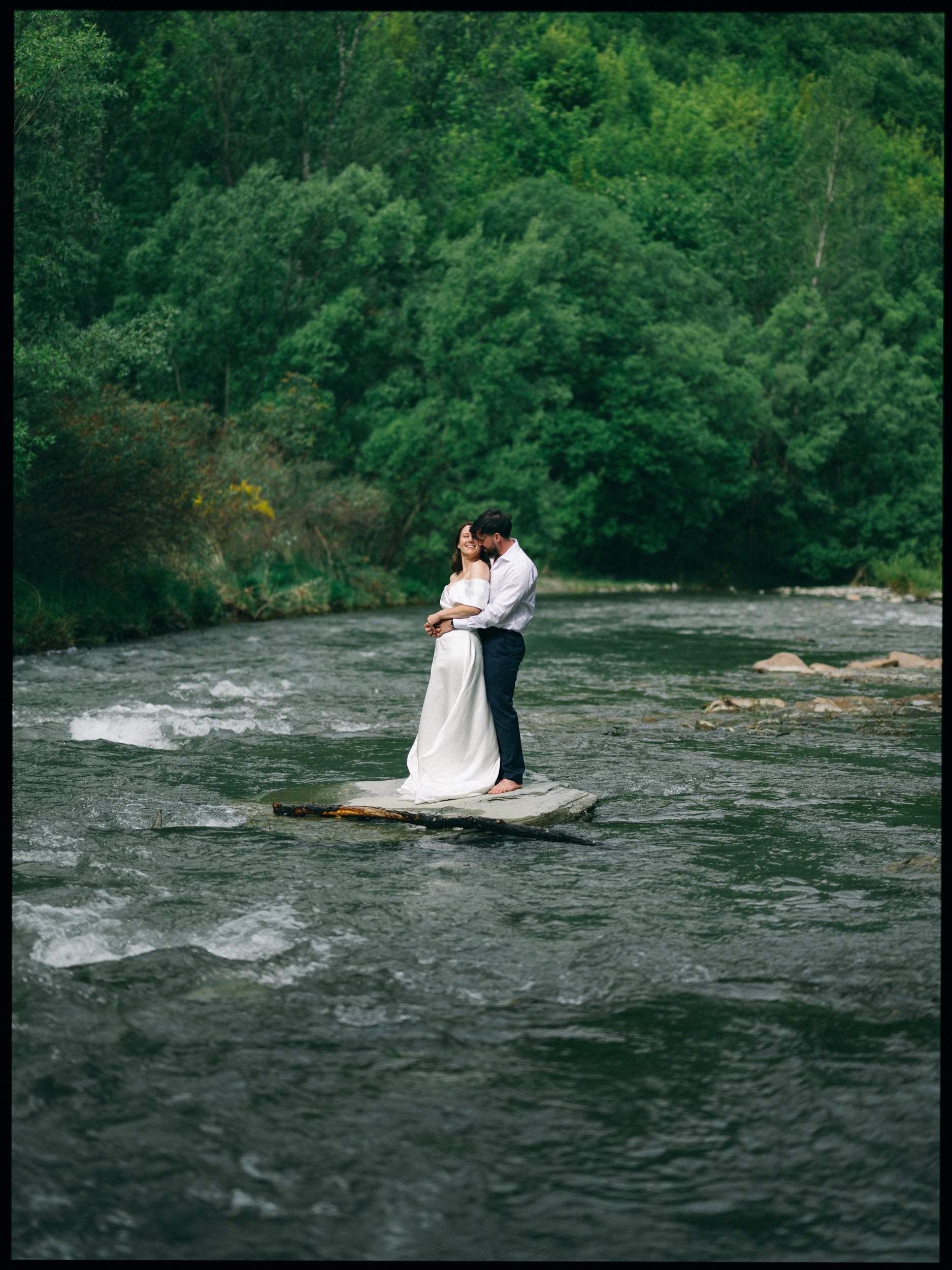 Queenstown Elopement, Otago, New Zealand. A couple in wedding attire standing and holding each other on a stone in the middle of the Arrow river in Arrowtown and surrounded by green trees.