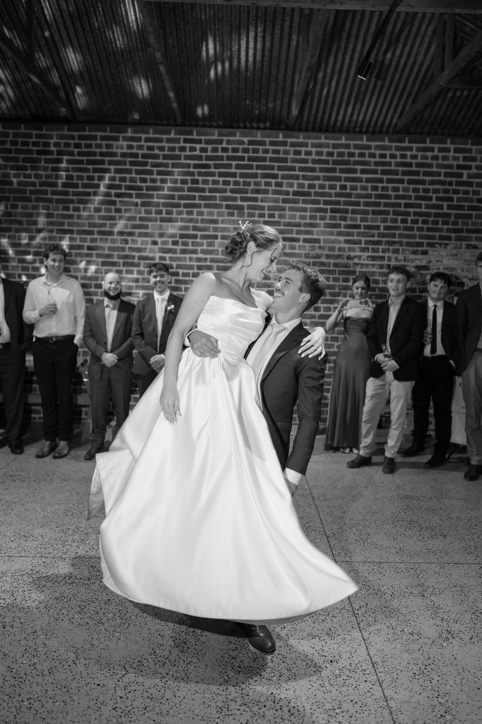 Film + Digital Wedding Photographer Dunedin - Lochend Woolshed, Dunedin, Otago, New Zealand. Bride and groom first dance at wedding reception, surrounded by friends and family against a brick wall background.