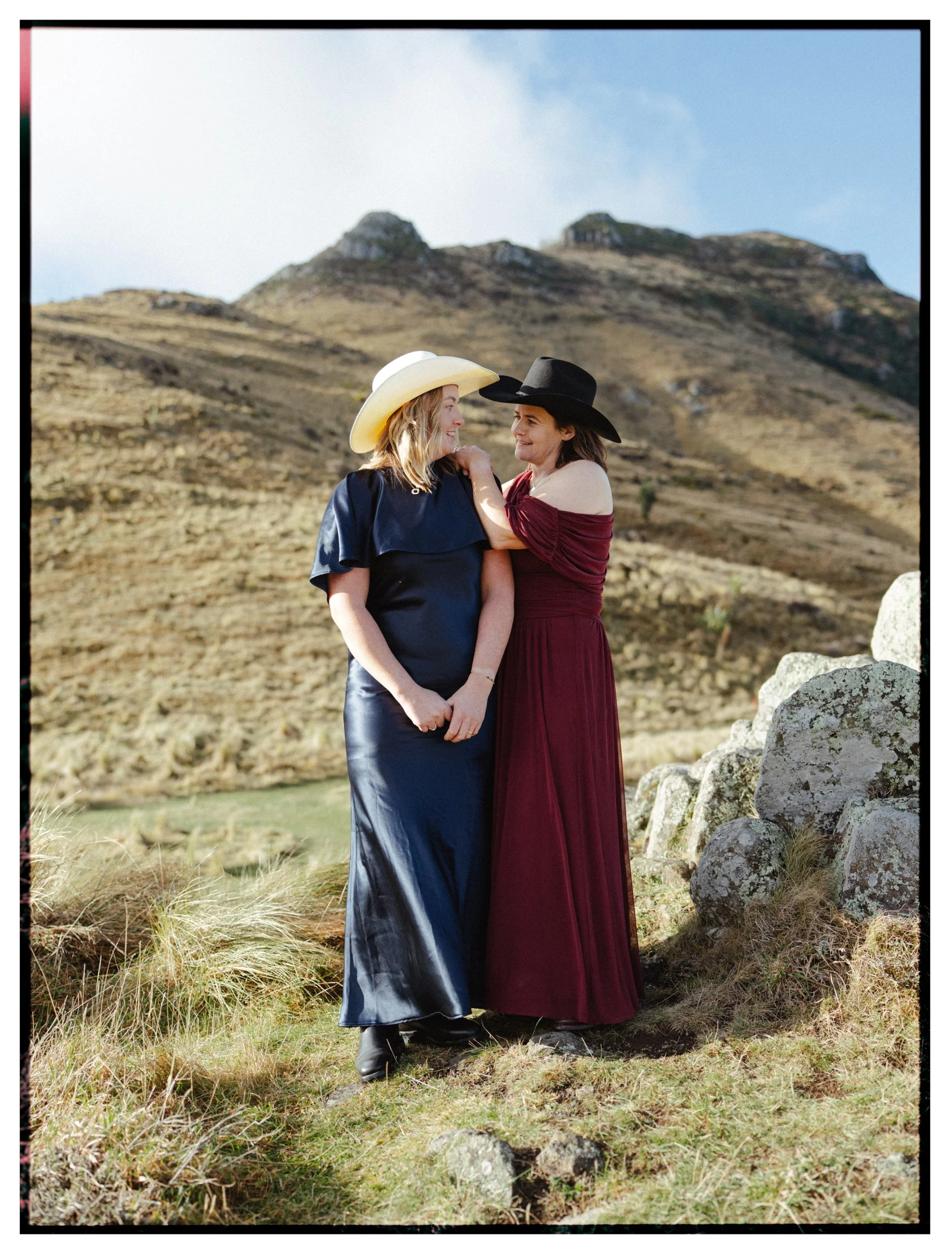 Film + Digital Wedding Photographer Christchurch - Banks Peninsula Wedding, Christchurch, New Zealand. Two brides standing on a grassy hillside, wearing cowboy hats, smiling and looking at each other, with mountains in the background.