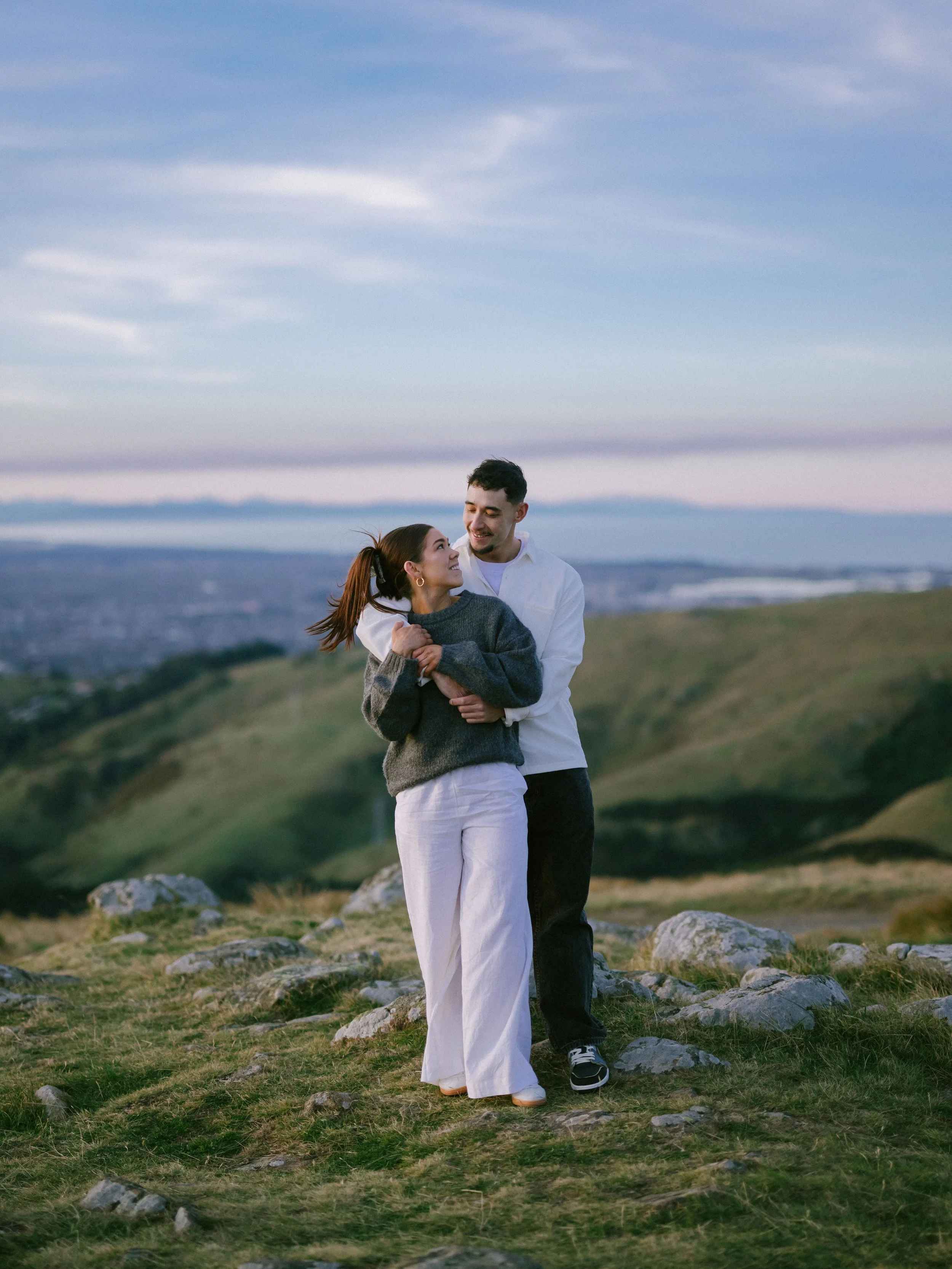 Fashionably dressed couple in loving embrace smiling at each other in the Port Hills above Cashmere in Christchurch, New Zealand with the ocean and Kaikoura mountains in the background.
