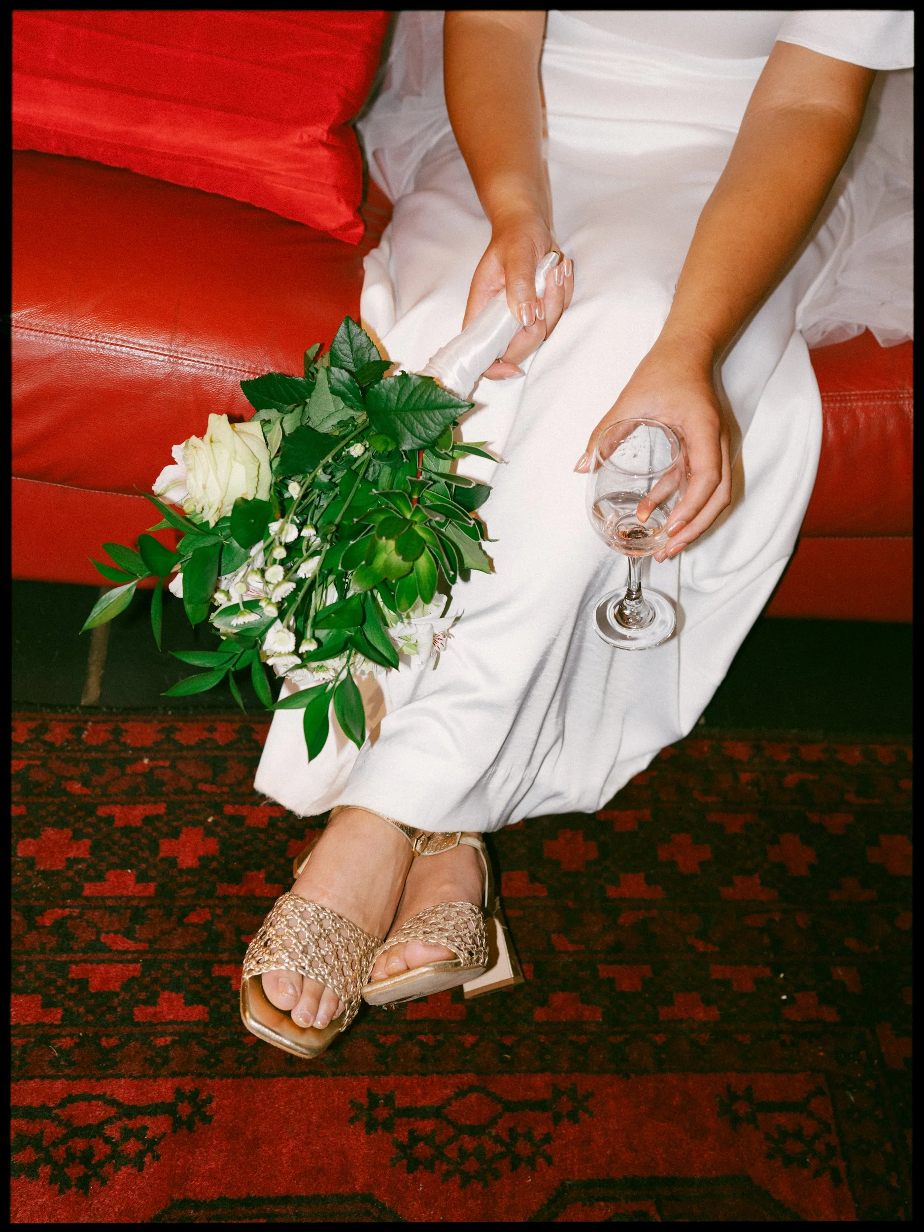 Film + Digital Wedding Photographer Christchurch - Backyard Wedding, Christchurch, Canterbury, New Zealand. Bride sitting on a red leather couch, holding a glass and bouquet of flowers. The floor has a red and black patterned carpet.
