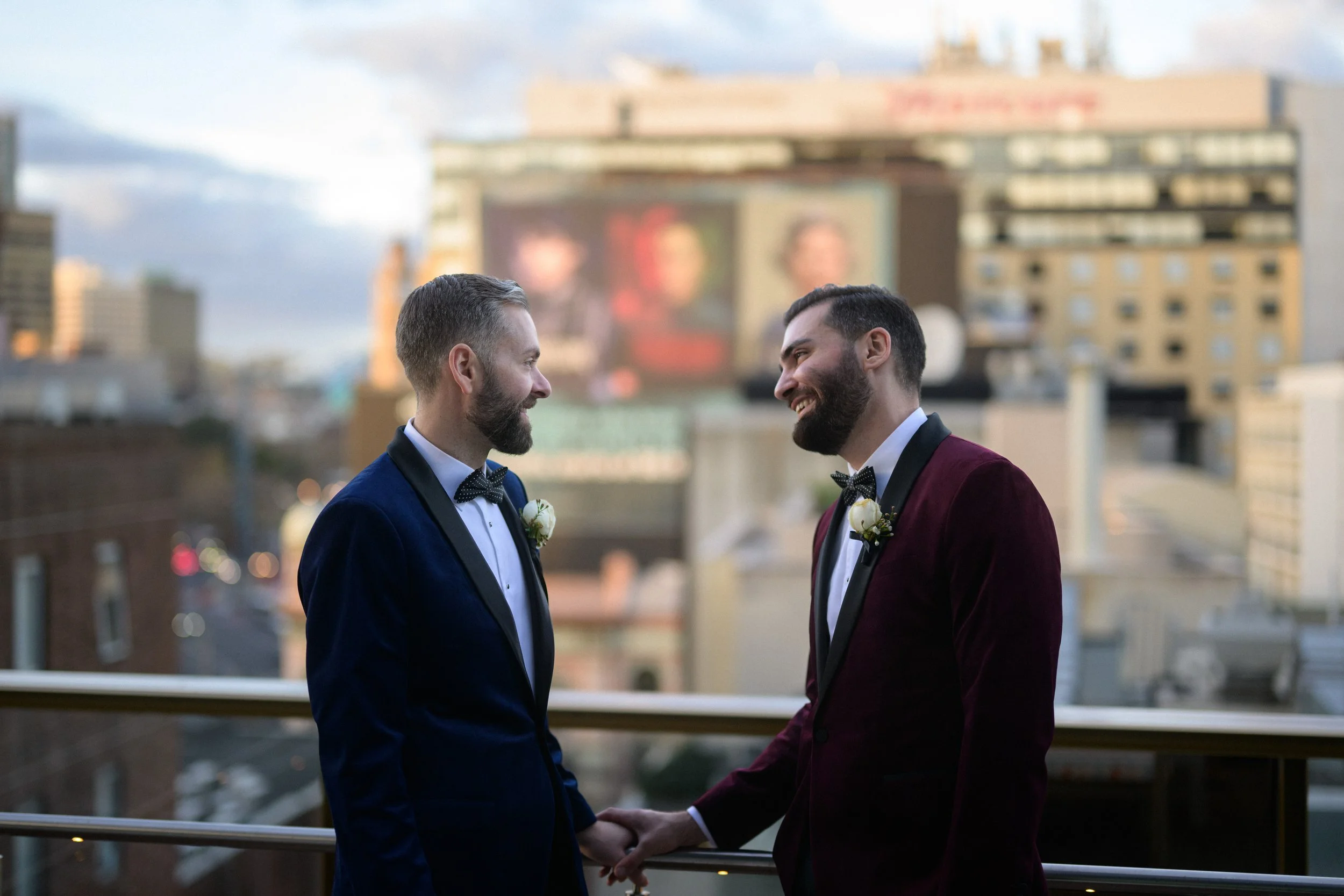 Grooms smiling at each other on hotel rooftop showing city landscape.