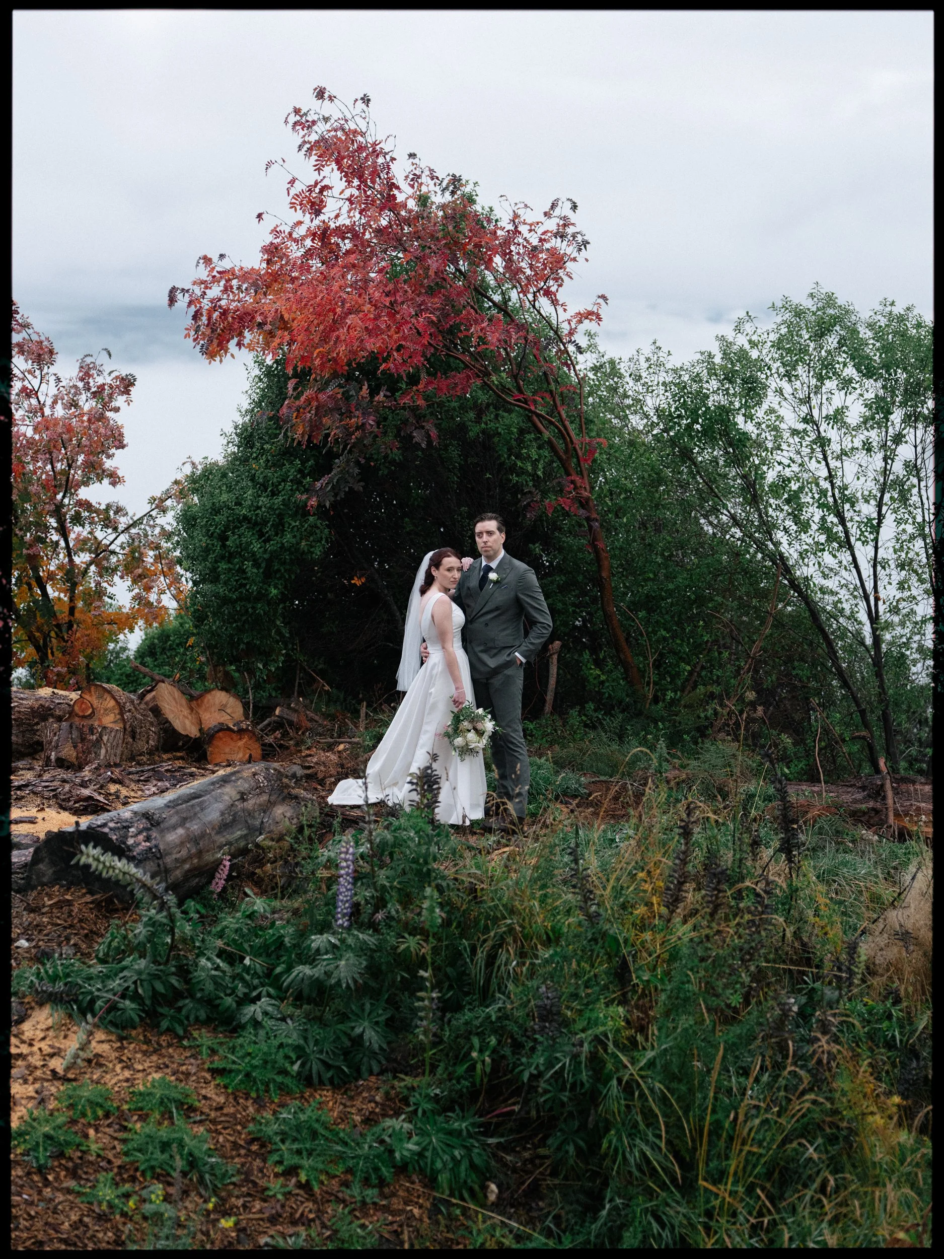 Film + Digital Wedding Photographer Queenstown - Kāmana Lakehouse, Queenstown, Otago, New Zealand. Bride and groom standing in a wooded area with colourful autumn trees and fallen logs near Moke Lake. Moody wedding photography.