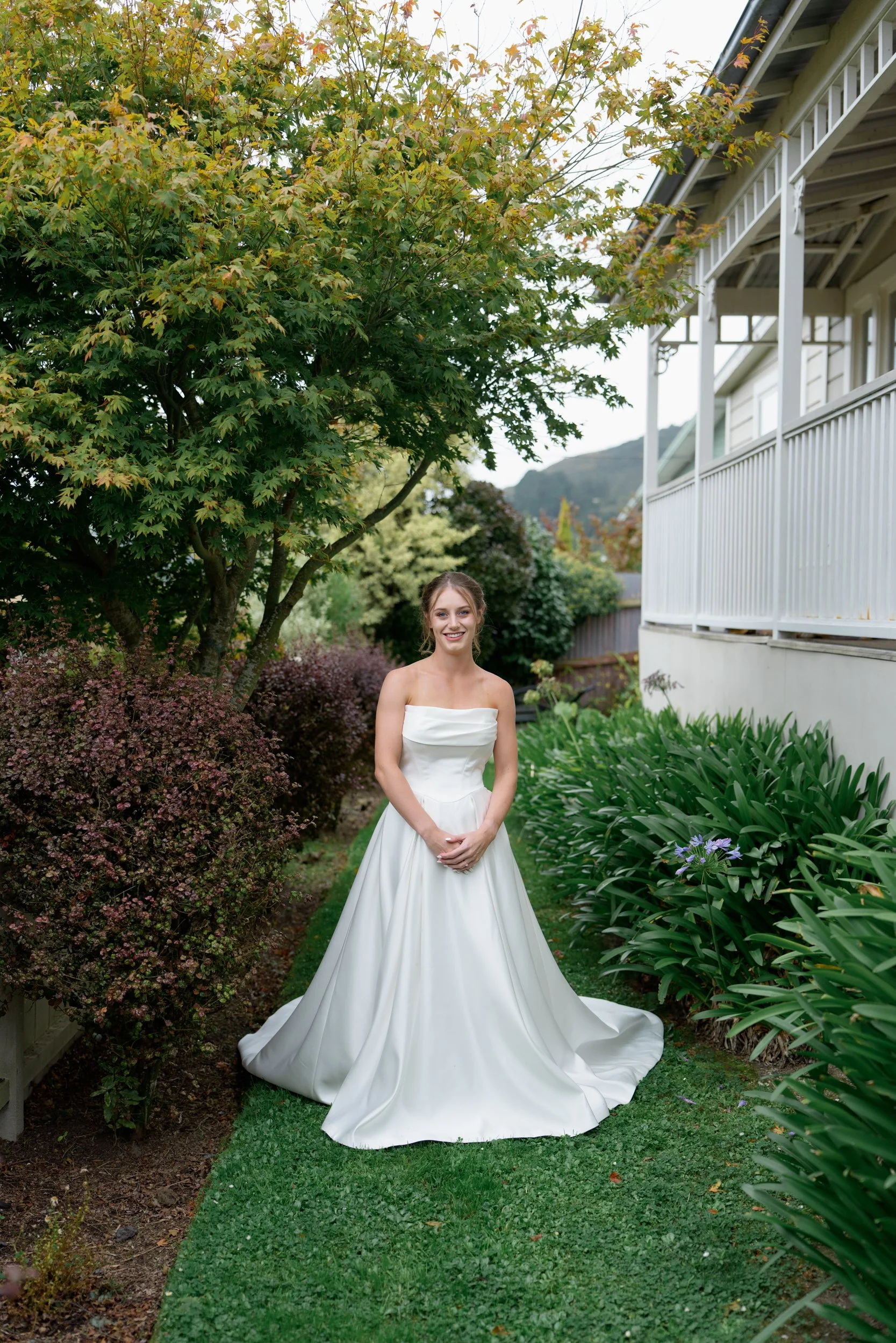 Film + Digital Wedding Photographer Dunedin - Lochend Woolshed, Dunedin, Otago, New Zealand. Bride in a white wedding gown standing in a garden with green plants and trees.