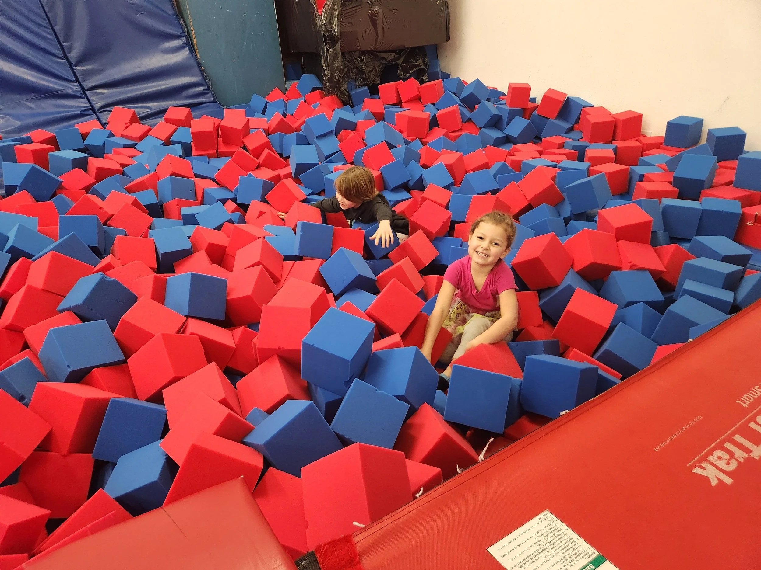 Young boy climbing into a large foam cylinder with supervision at indoor gymnastics or play area.