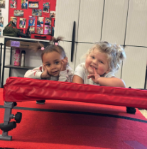 Two young girls lying on a red mat, smiling and resting their heads on their hands, in a room with white panels and shelves in the background.