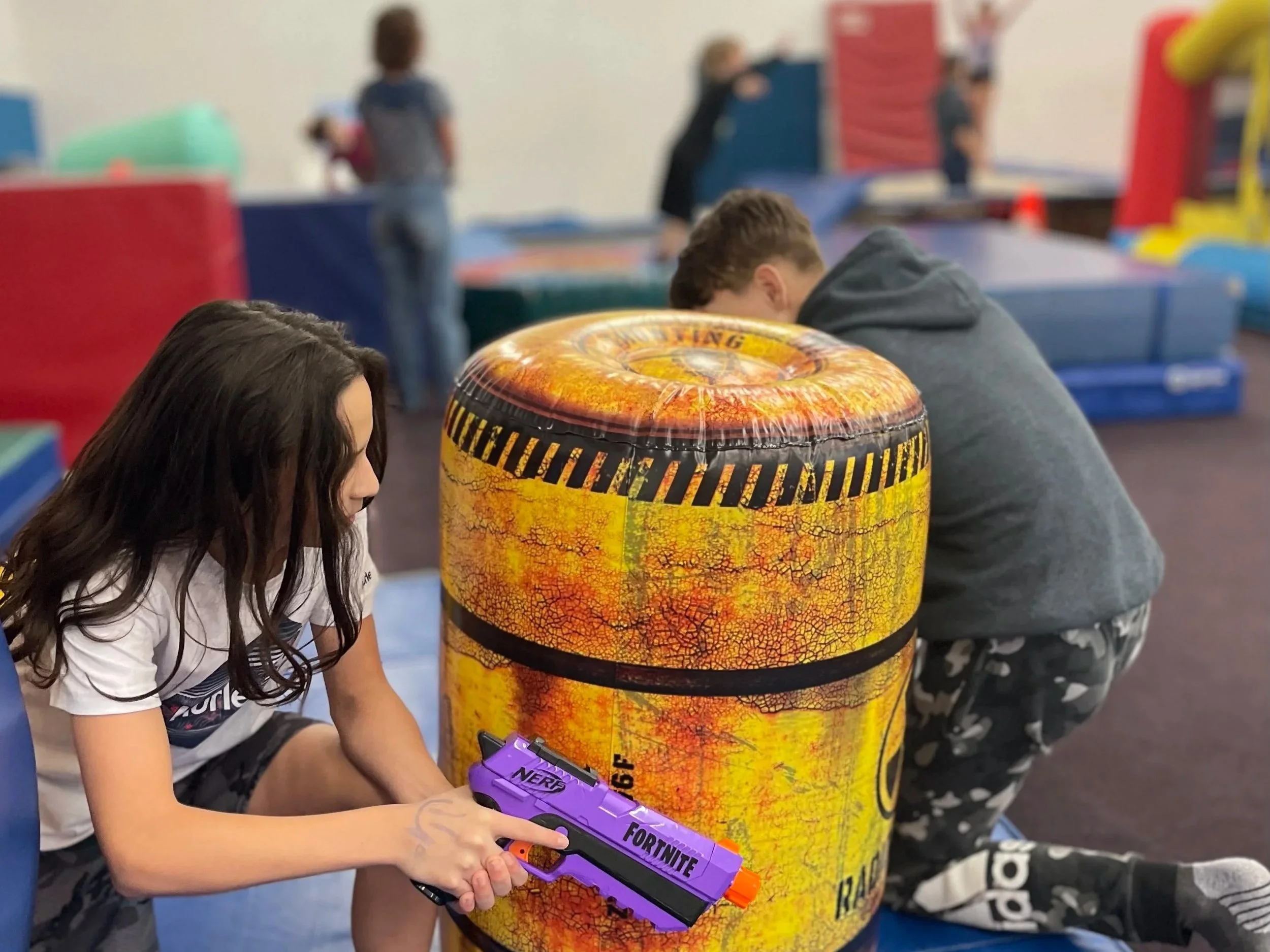 Children playing in an indoor gymnastics gym, with various colorful foam mats, balance beams, and gymnastic equipment, under a high ceiling with exposed beams.
