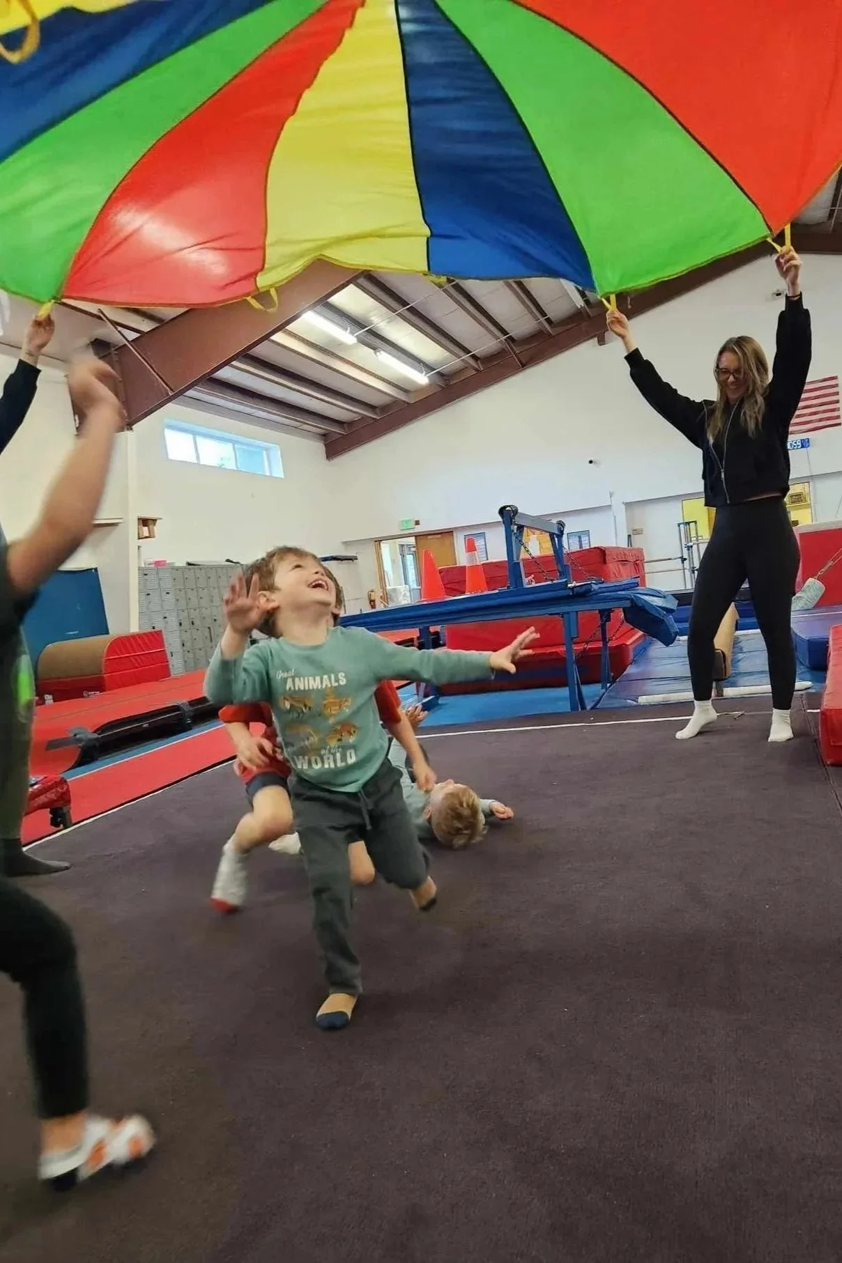 Children playing with a woman under a large colorful parachute in an indoor gym or gymnastics facility.