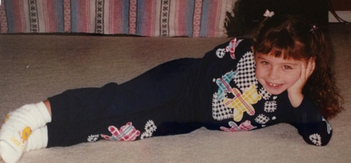 A picture of Mandy when she was a child. She is laying on the ground, leaning on her hand, smiling at the camera.