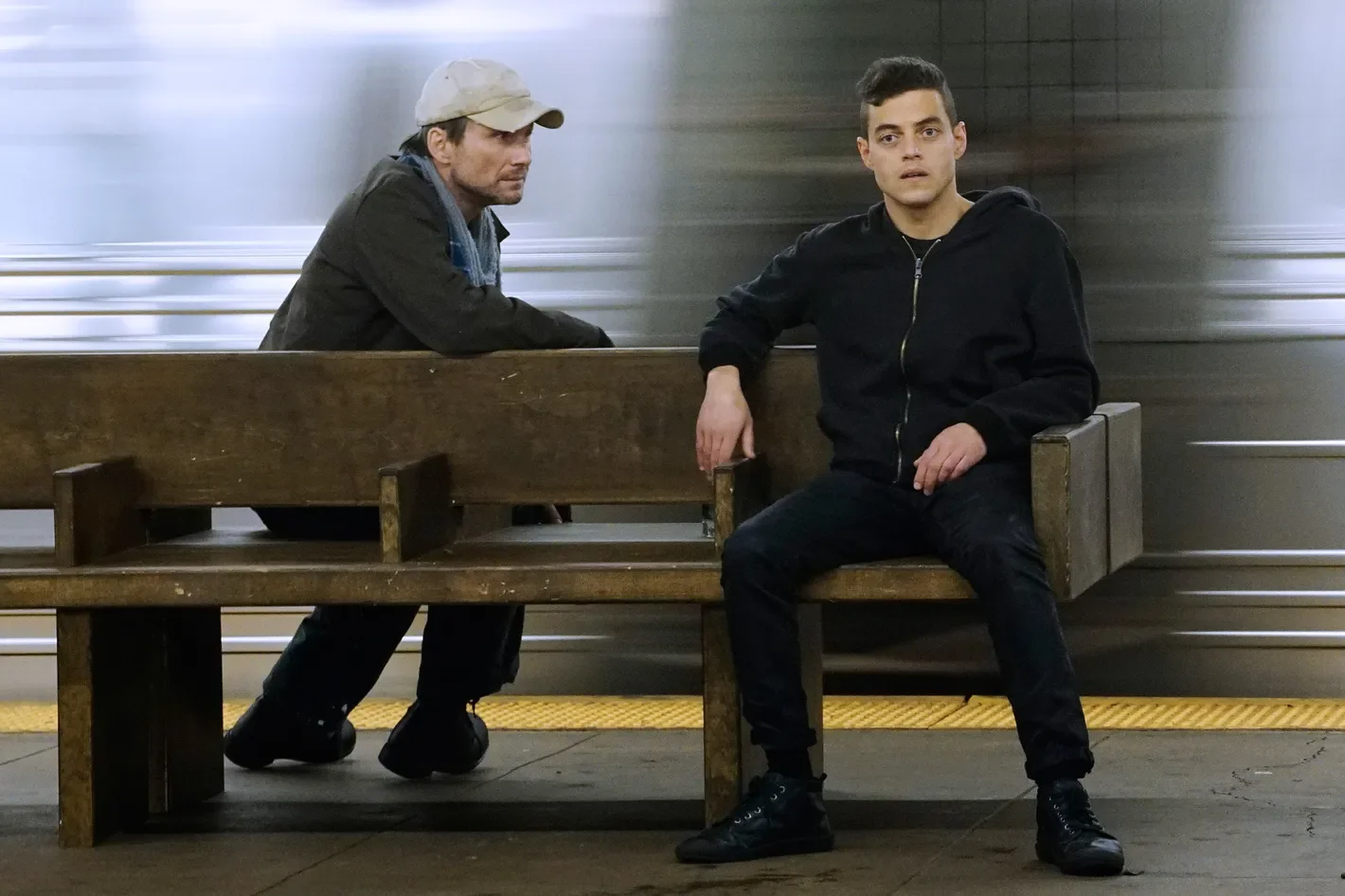 Elliot sitting on a bend, and Mr. Robot looking at Elliot from the other side of the bench